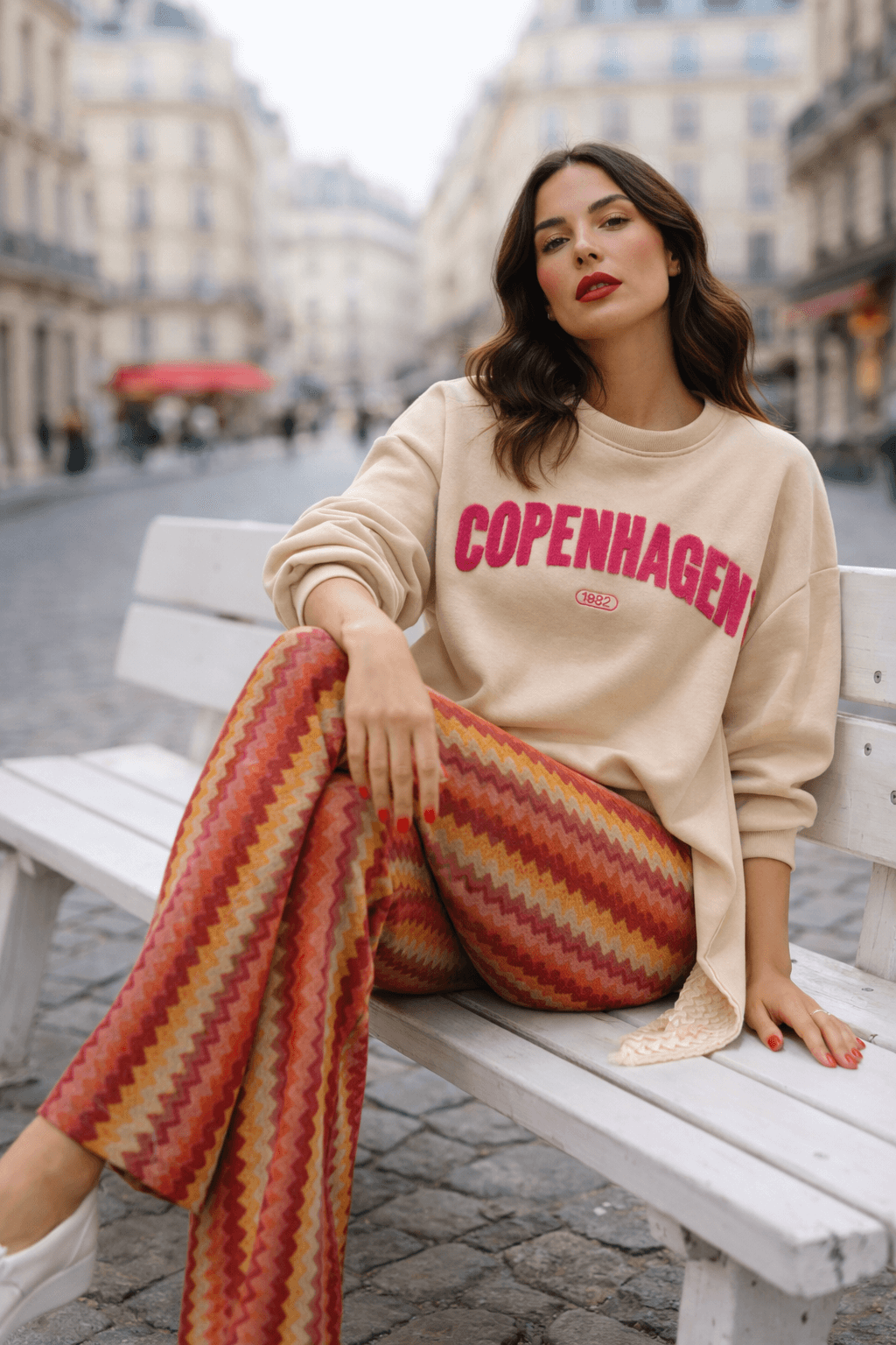 Frau sitzt auf einer weißen Holzbank in einer Pariser Innenstadtstraße mit Kopfsteinpflaster und unscharfen Altbauten im Hintergrund. Sie trägt ein cremefarbenes Sweatshirt mit pinkem Schriftzug „COPENHAGEN 1982“ sowie eine weite Hose mit Zickzackmuster in Rot-, Orange- und Gelbtönen.
