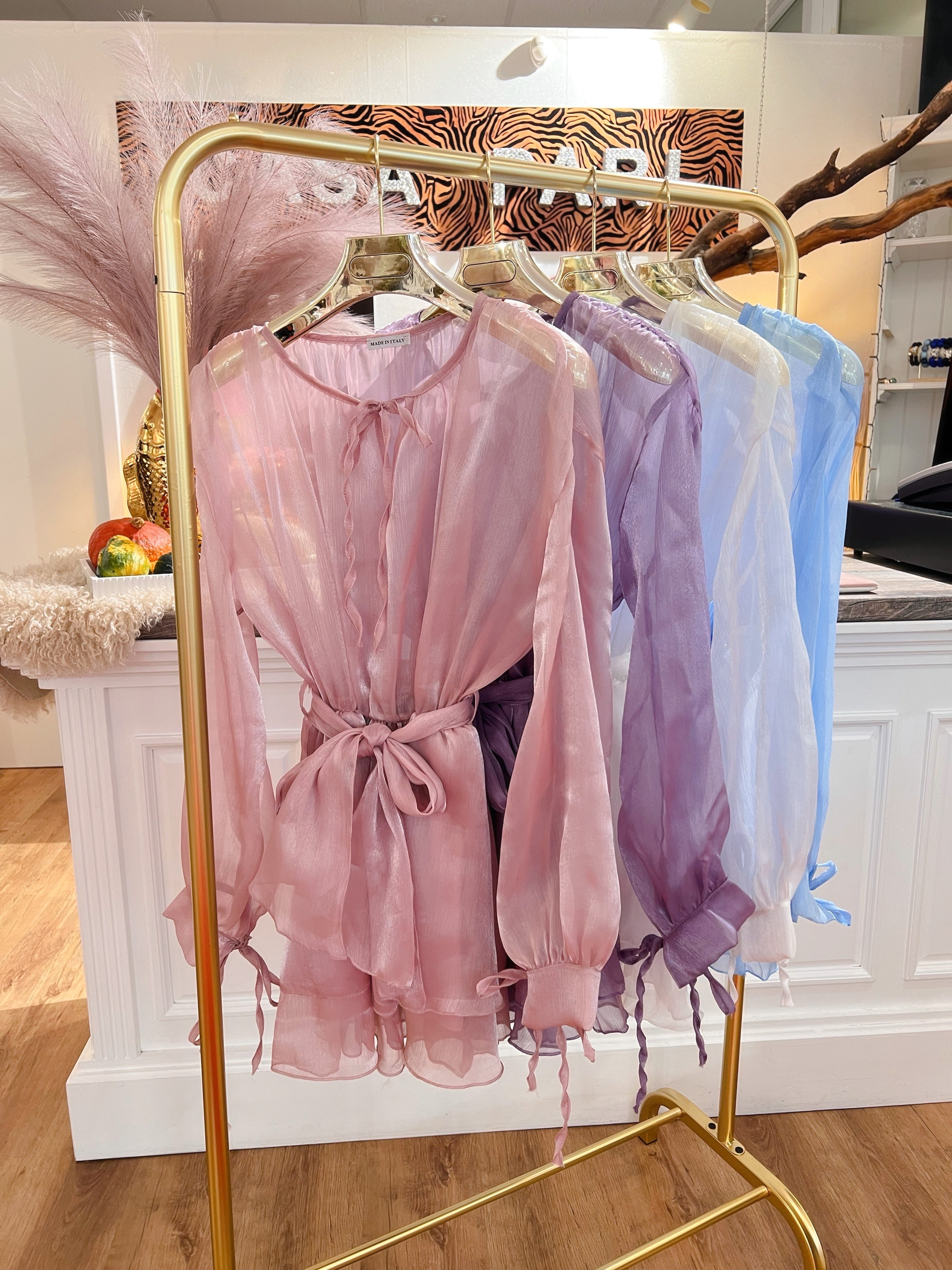 Mehrere transparente Blusen in Rosa, Flieder, Weiß und hellblau hängen auf goldenen Kleiderbügeln an einem goldenen Kleiderständer vor weißen Theke im Laden.