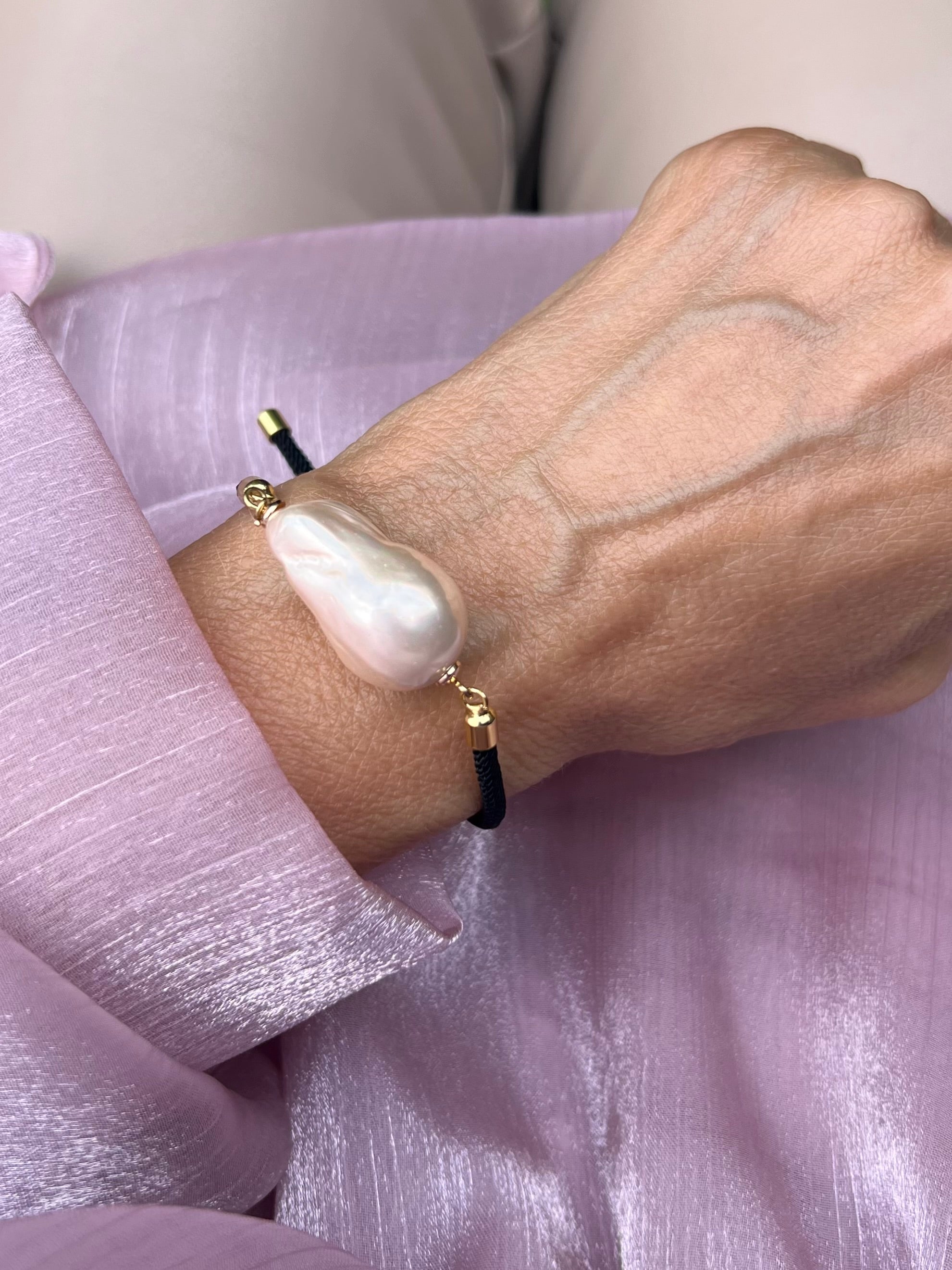 Handgelenk mit feinem schwarzen Armband und einer Barockperle mit goldenen Details. Auf rosa Stoff fotografiert.