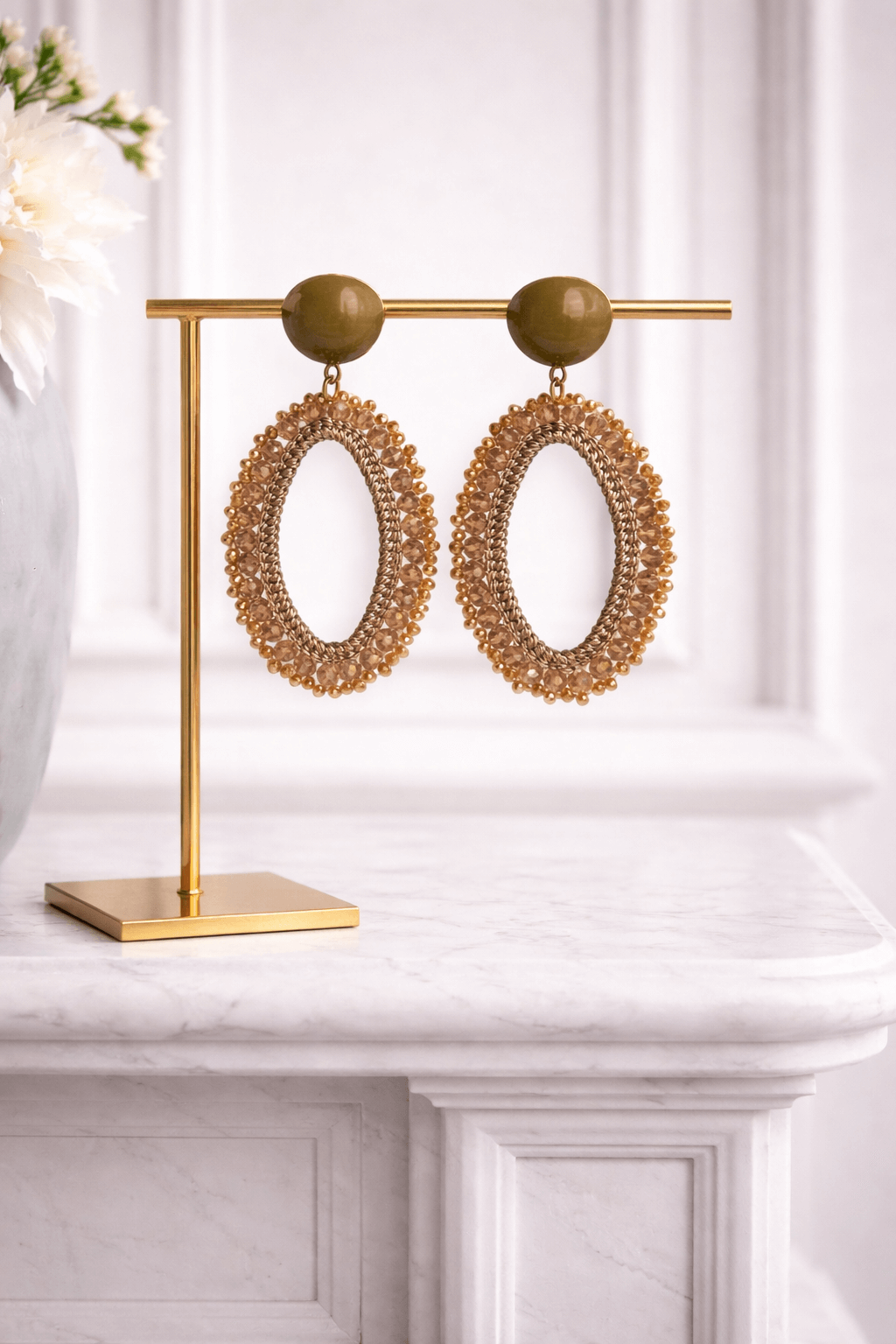 Ovale Ohrringe mit beigefarbenem Stecker, goldfarbenem Garnrahmen und umlaufenden champagnerfarbenen Glasperlen, präsentiert auf einem goldfarbenen Schmuckständer vor hellem Hintergrund.