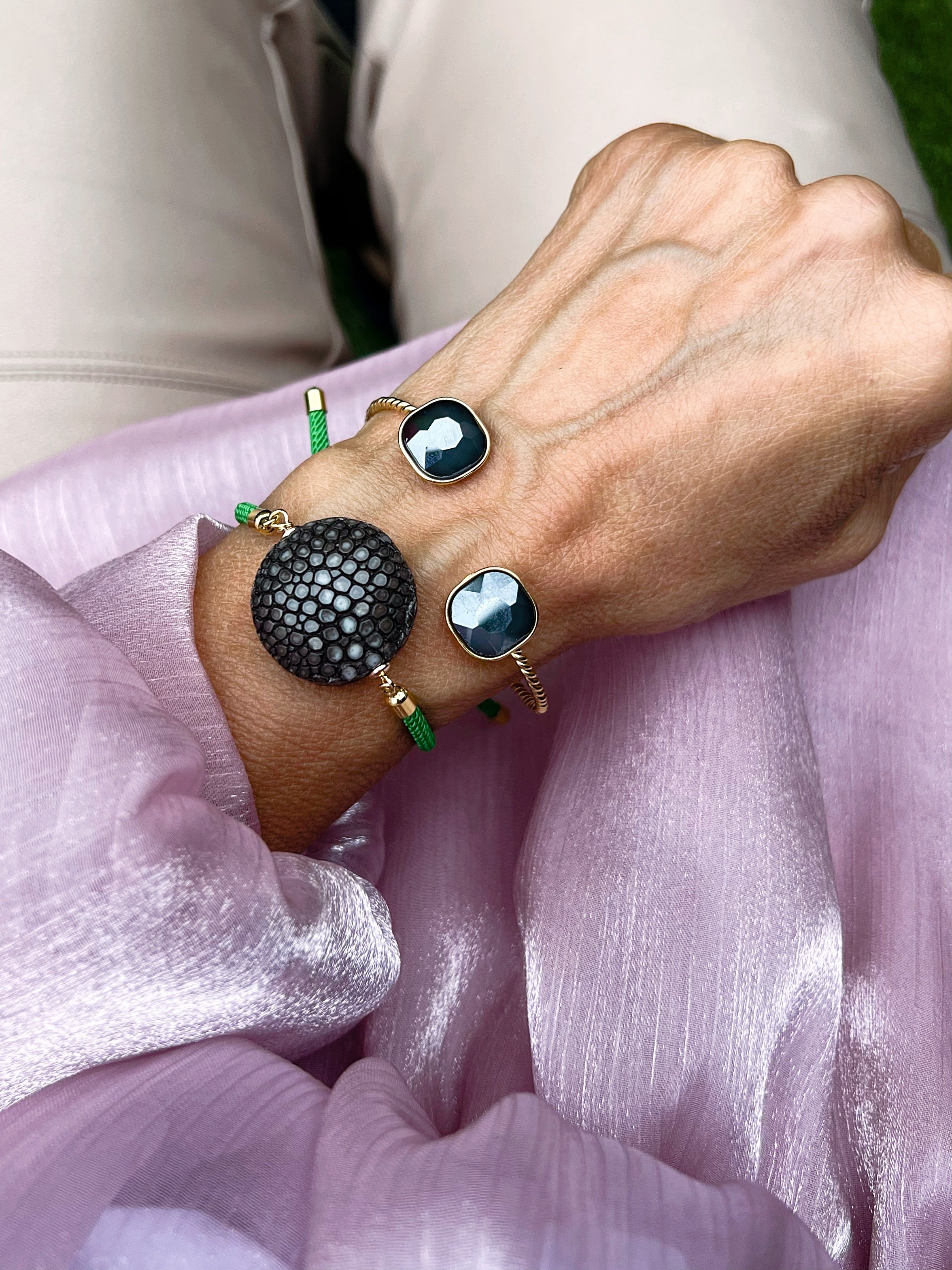 Ein grünes Armband mit schwarzer Perle dazu ein goldener Armreif mit Glasstein in Taupe. An einem Handgelenk auf rosa Stoff fotografiert.