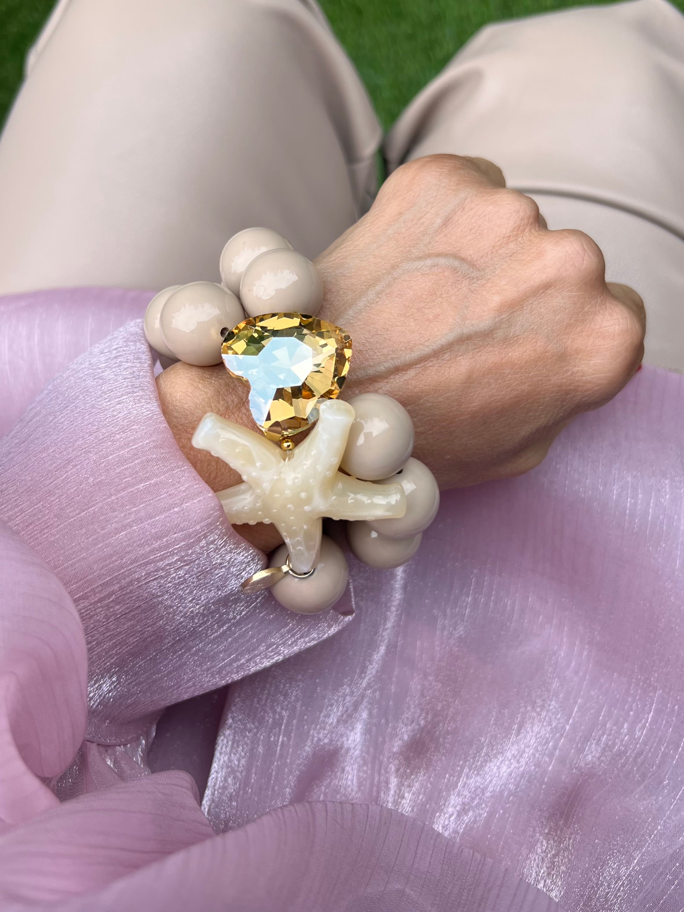 Armband mit zwei Reihen glänzender Perlen in Beige, verziert mit einem funkelndem Kristalle Herz in Champagner, auf hellem Hintergrund.