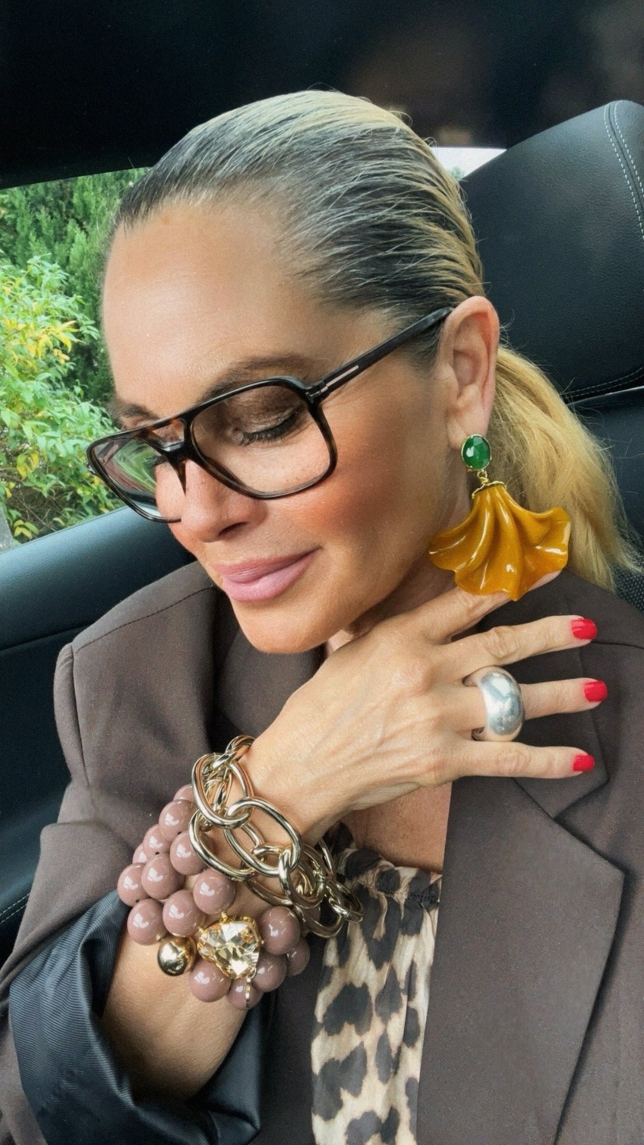 Frau sitzt im Auto und trägt einen großen, goldgelben Fächer-Ohrring mit grünem Jadestecker. Sie hält eine Hand unter das Ohr, um den Ohrring zu betonen. Dazu trägt sie ein braunes Sakko, goldfarbenen Armreif und ein Armband aus taupefarbenen Perlen.