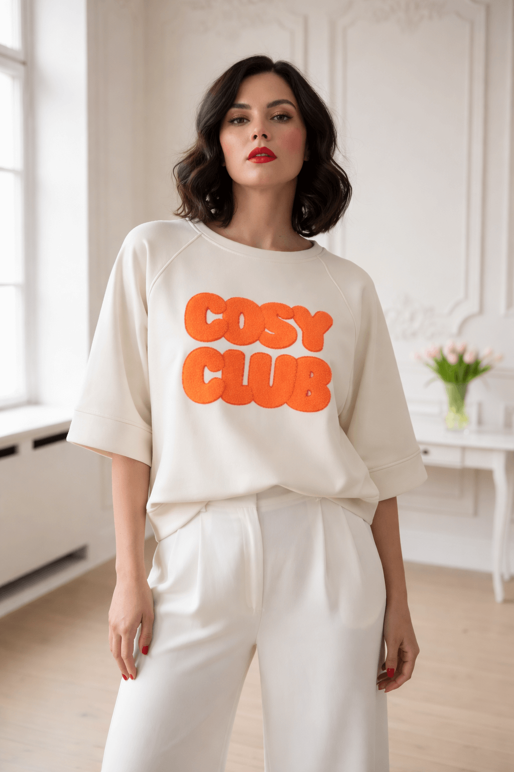 Frau mit dunkelbraunem, schulterlangem Haar trägt ein helles Sweatshirt mit orabgem „Cosy Club“ Frottee-Schriftzug, kombiniert mit weißer, weiter Hose, in hellem Innenraum.