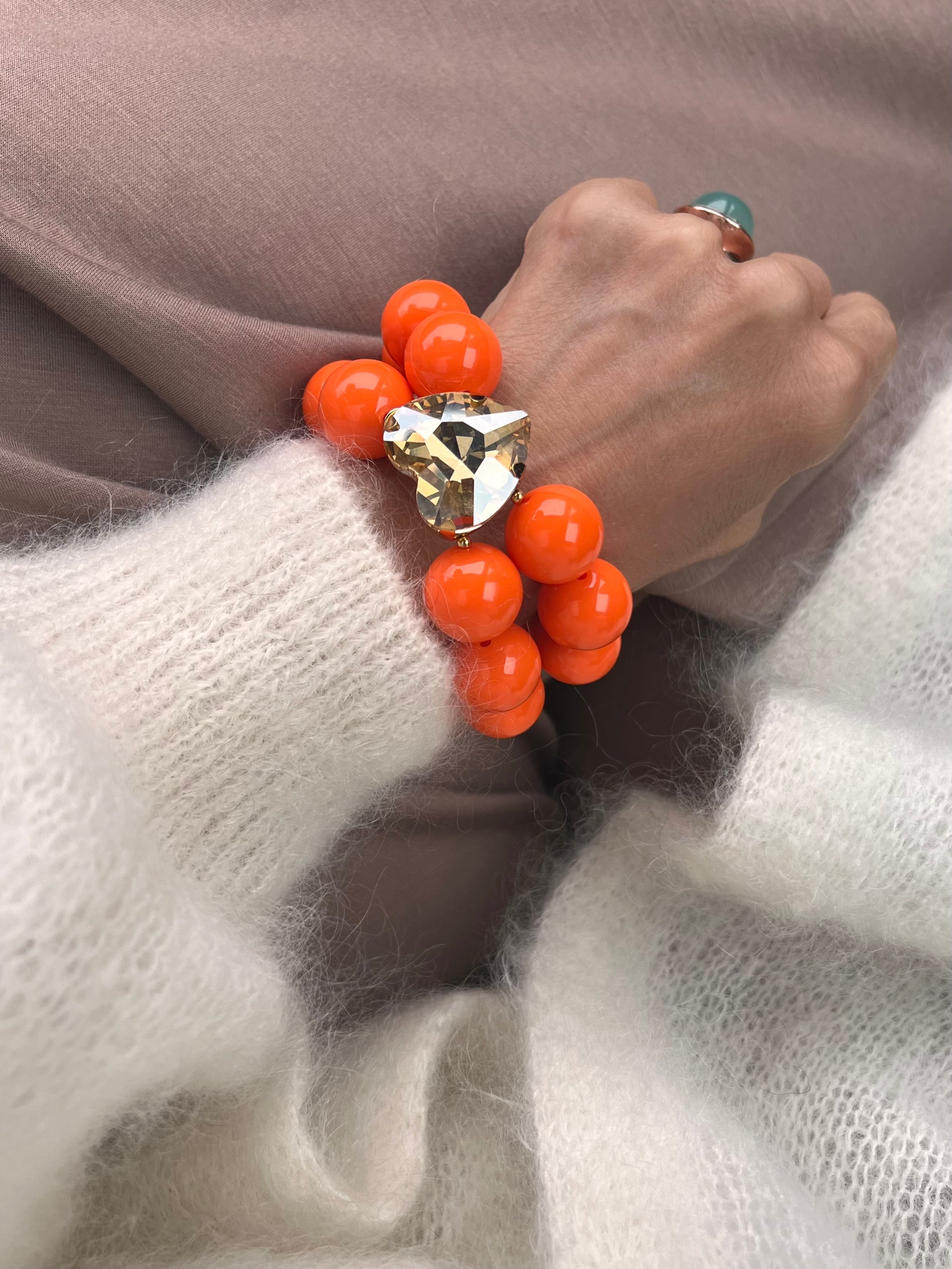 Armband in Orange mit großem Herzschmuckstein, und großem Ring. Am Handgelenk auf beigem Stoff fotografiert.