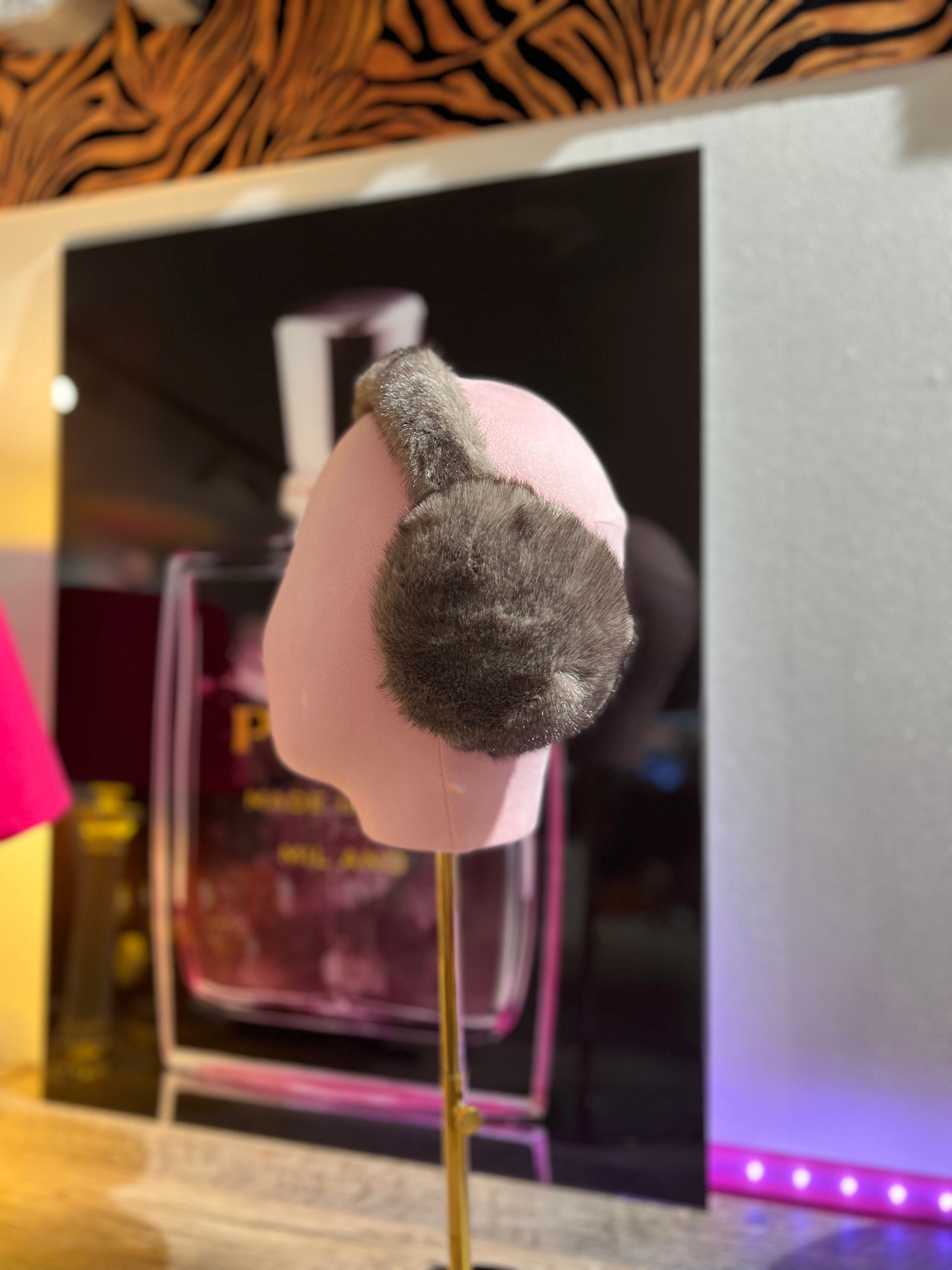 Taupe, flauschige Ohrwärmer auf rosafarbene Präsentation Büste, im Hintergrund unscharf ein Parfumflakon und warme Beleuchtung.