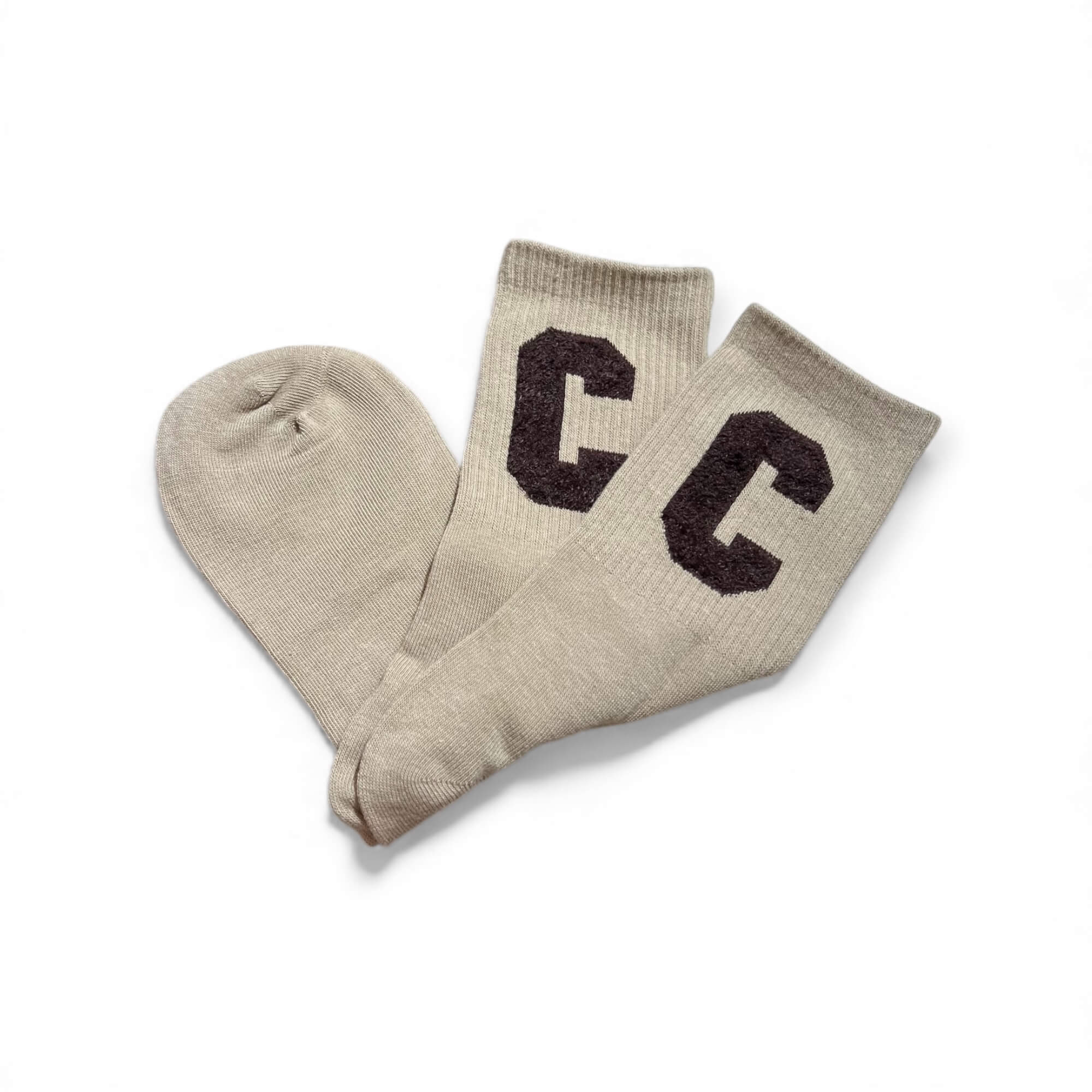 Beige kurze Socken aus weichem Strick, flach liegend fotografiert, mit zwei braunen „C“-Buchstaben auf dem Schaft. Die Socken liegen leicht überkreuzt auf weißem Hintergrund.