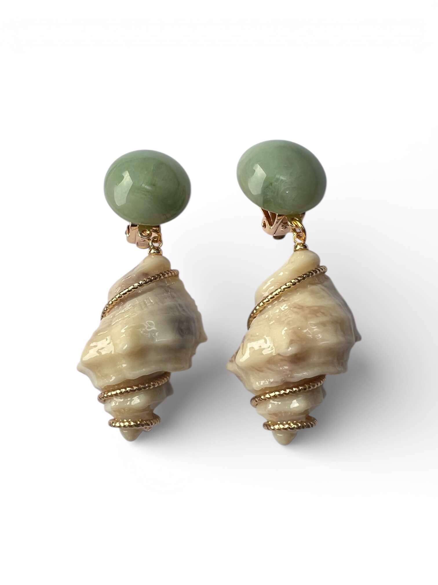 Paar grüne Clip-Ohrringe mit rundem Stein oben und cremefarbener Muschelform als Anhänger, verziert mit goldfarbenen Drahtdetails, freigestellt vor weißem Hintergrund.