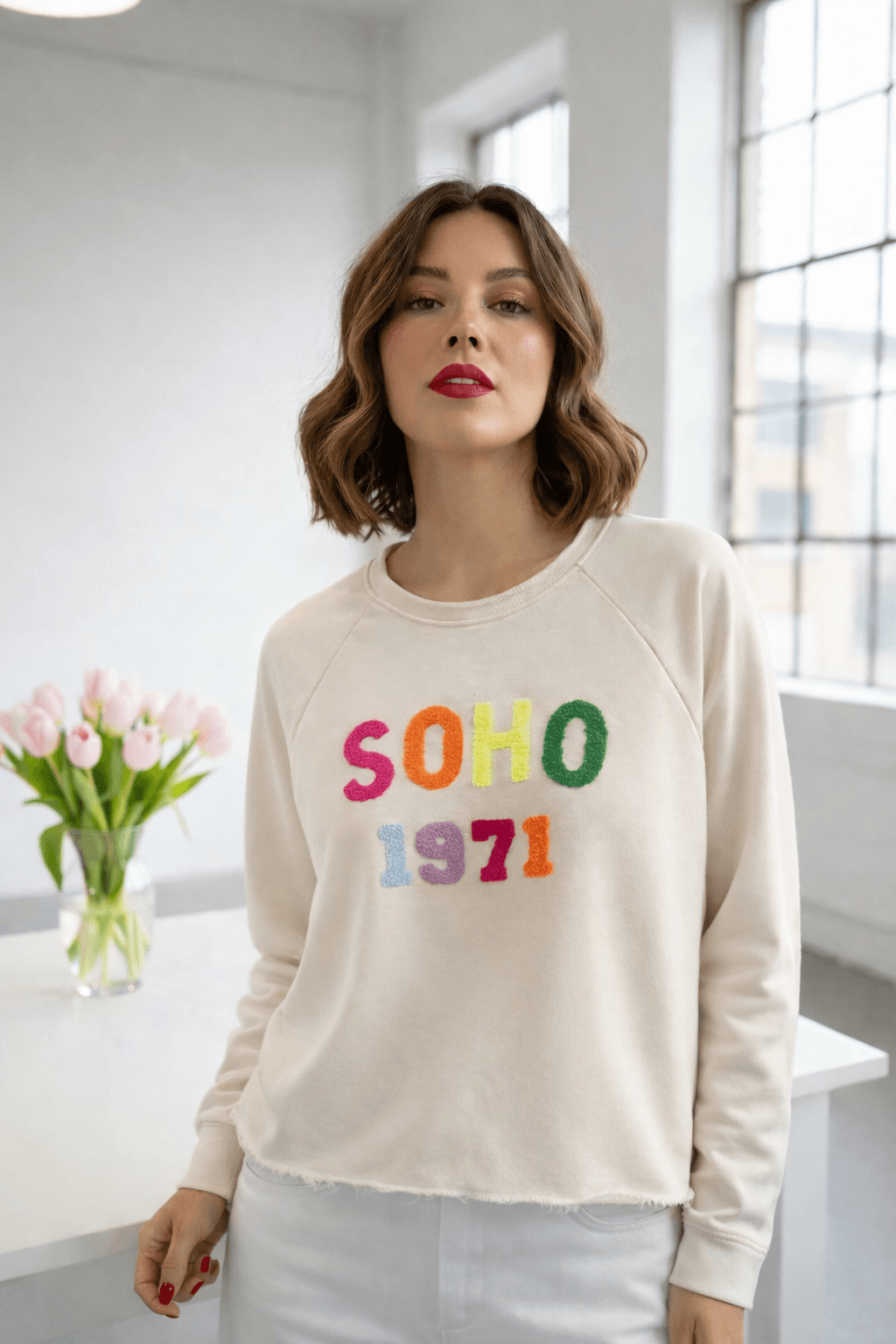 Frau trägt beigen SOHO 1971 Sweater mit buntem Schriftzug, lockerer Schnitt mit offenem Used-Look-Saum, kombiniert mit weißer Hose in hellem Raum mit Fenster und Tulpen.