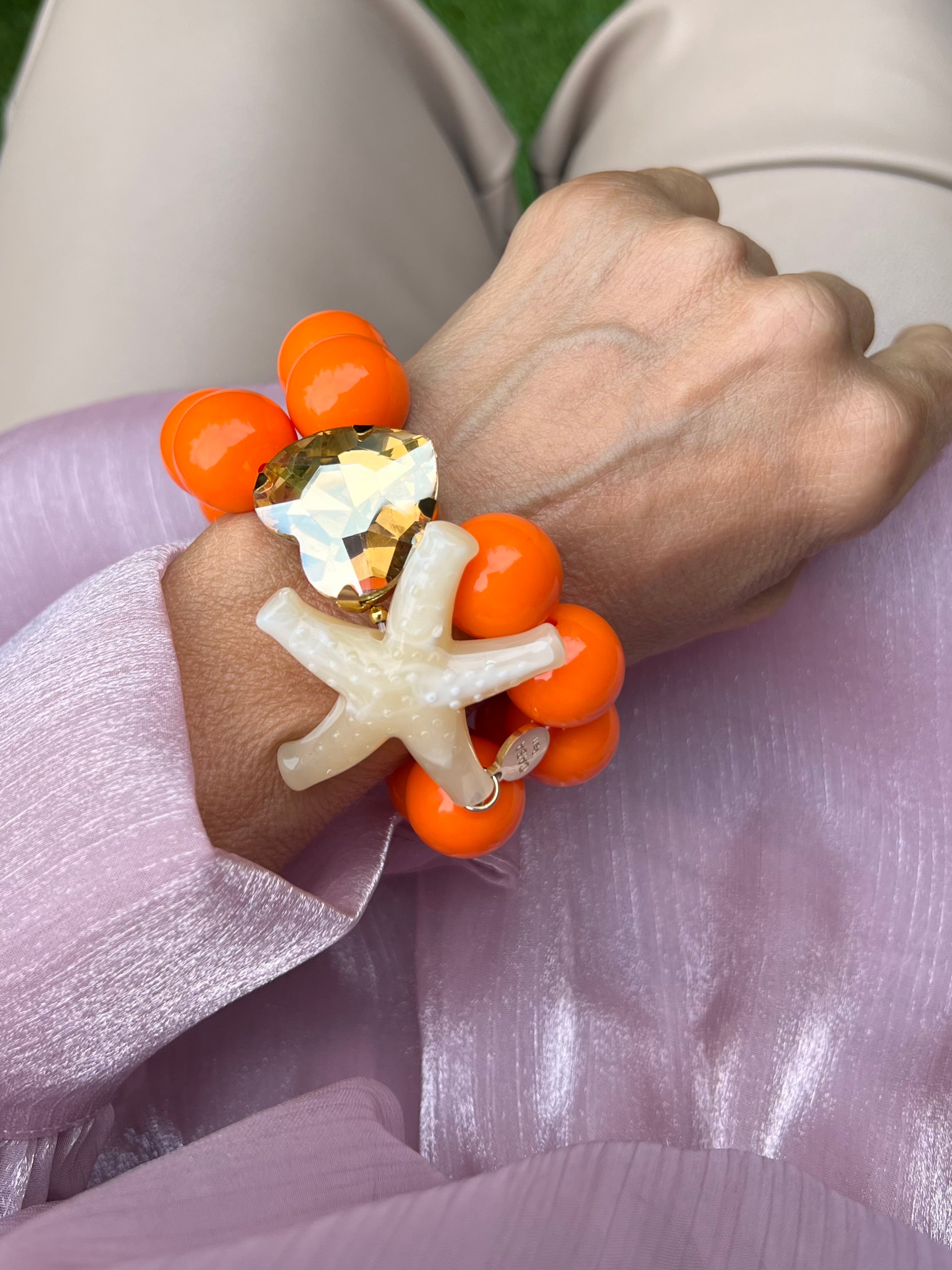 Armband mit zwei Reihen glänzender orangefarbener Perlen, verziert mit einem funkelndem Kristalle Herz in Champagner, auf hellem Hintergrund.