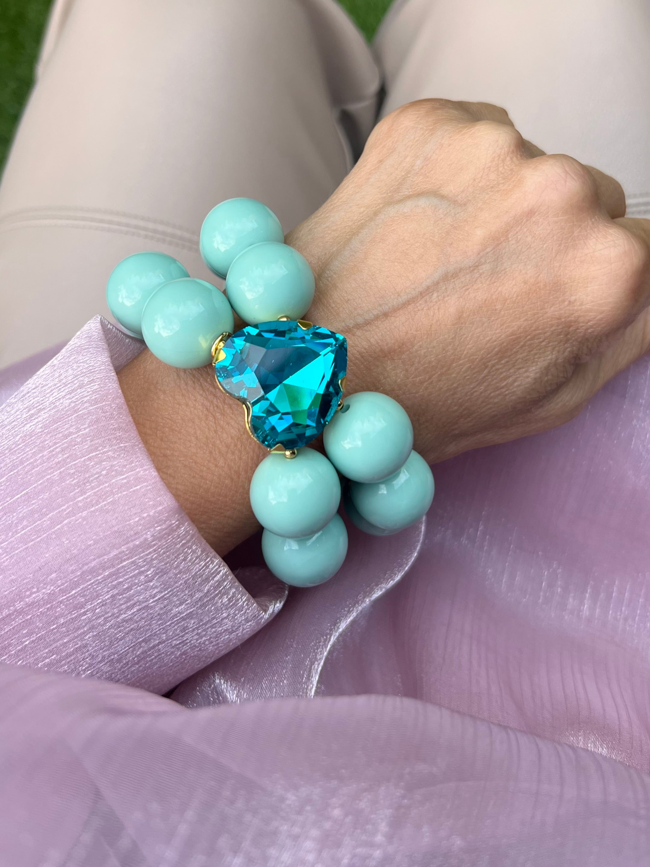 Handgelenk mit hell türkisem Armband aus großen, glänzenden Perlen und einem herzförmigen, tiefblauen Kristall in der Mitte.