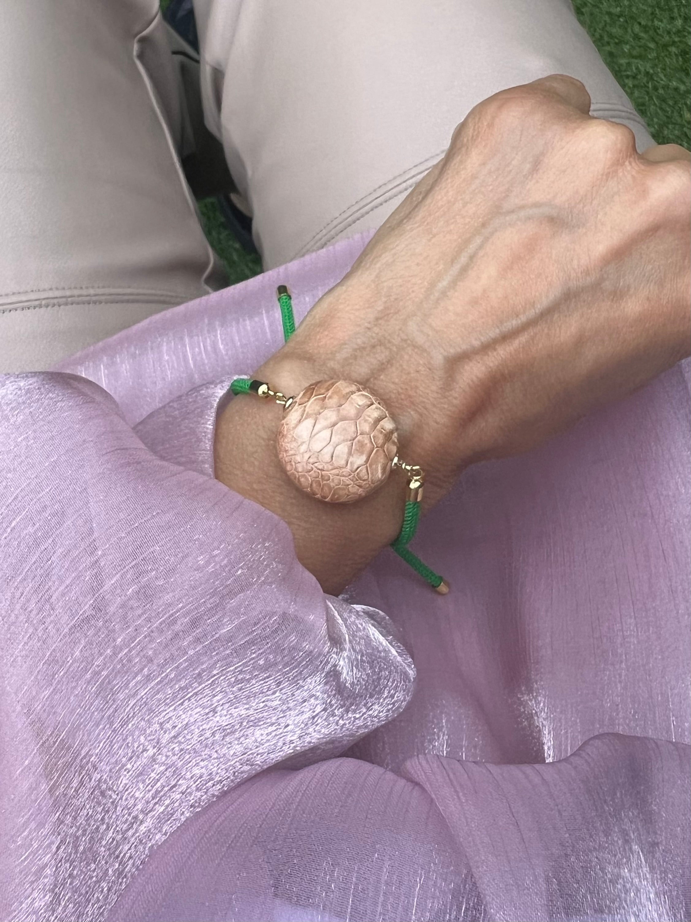 Grünes Armband mit beiger Perlen und goldenen Details. Fotografiert am Handgelenk auf rosa Stoff.