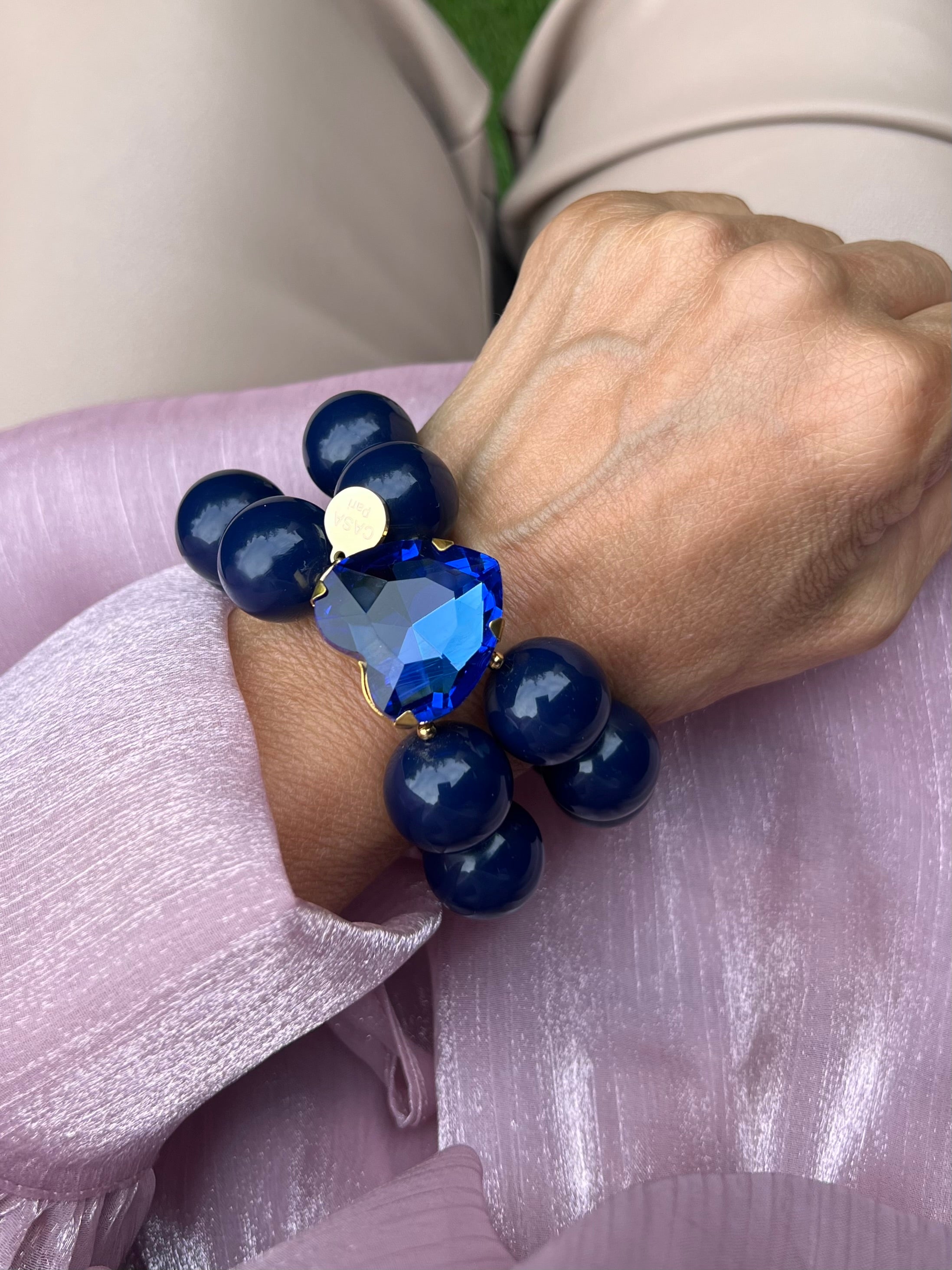 Handgelenk mit blauem Armband aus großen, glänzenden Perlen und einem herzförmigen, tiefblauen Kristall in der Mitte.