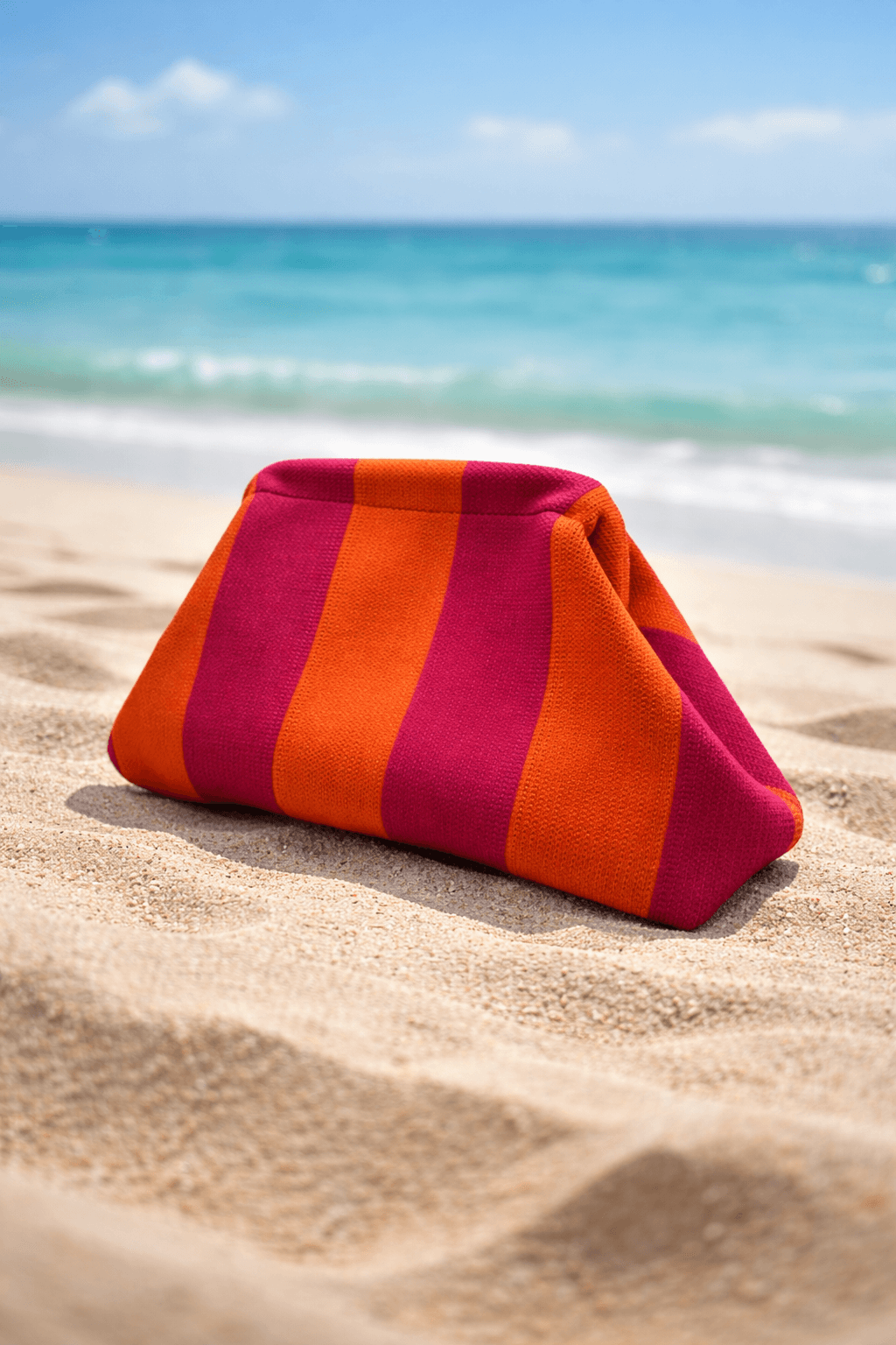 Gestreifte Clutch aus festem Canvas in Pink und Orange steht im Sand am Strand, im Hintergrund sind Meer und Himmel leicht unscharf zu sehen.