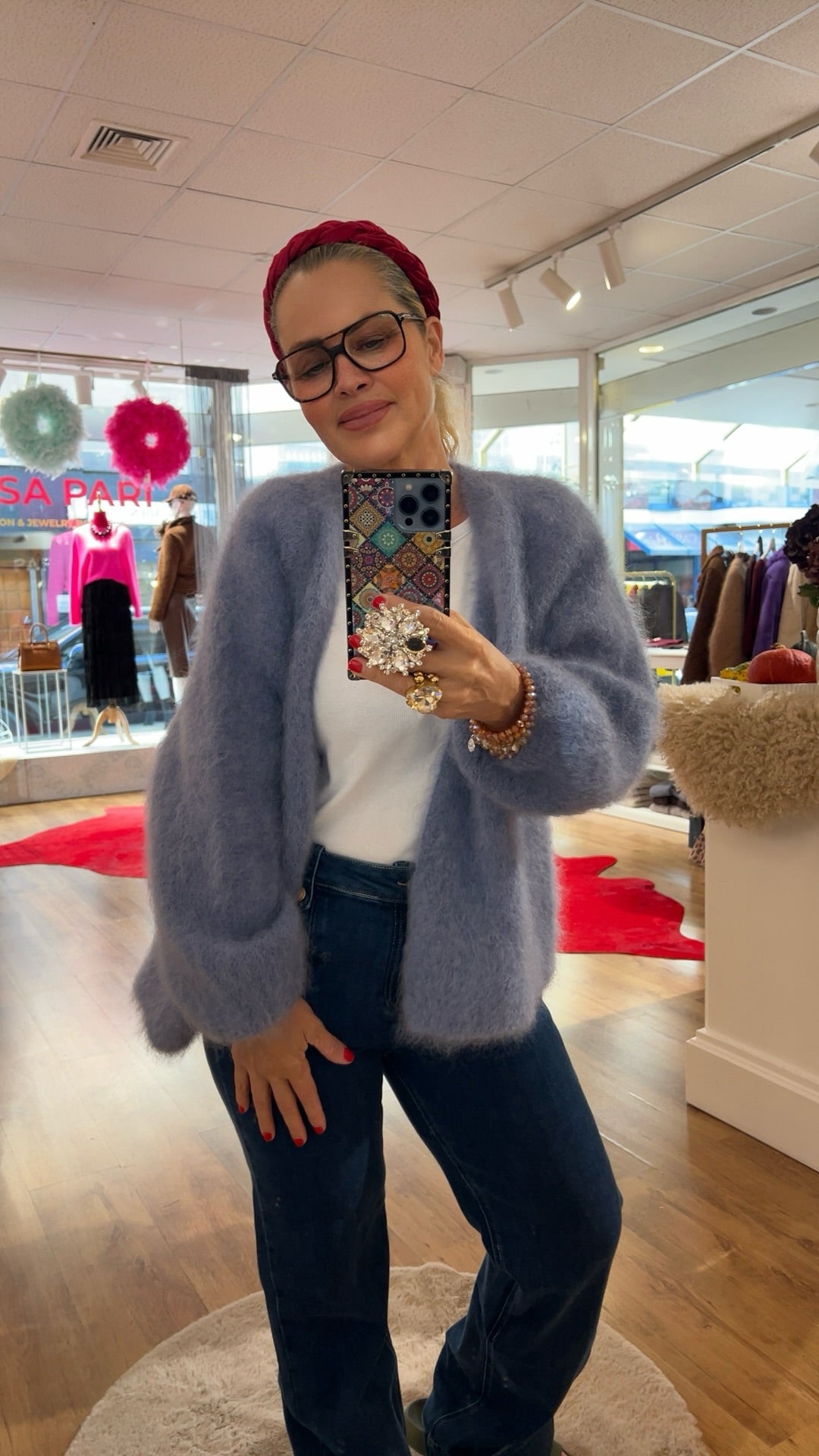 Frau trägt flauschige Strickjacke in Taubenblau, kombiniert mit weißem Shirt und dunkelblauer Jeans mit Knopfleiste. Im Hintergrund bunte Mode und rote Akzente.