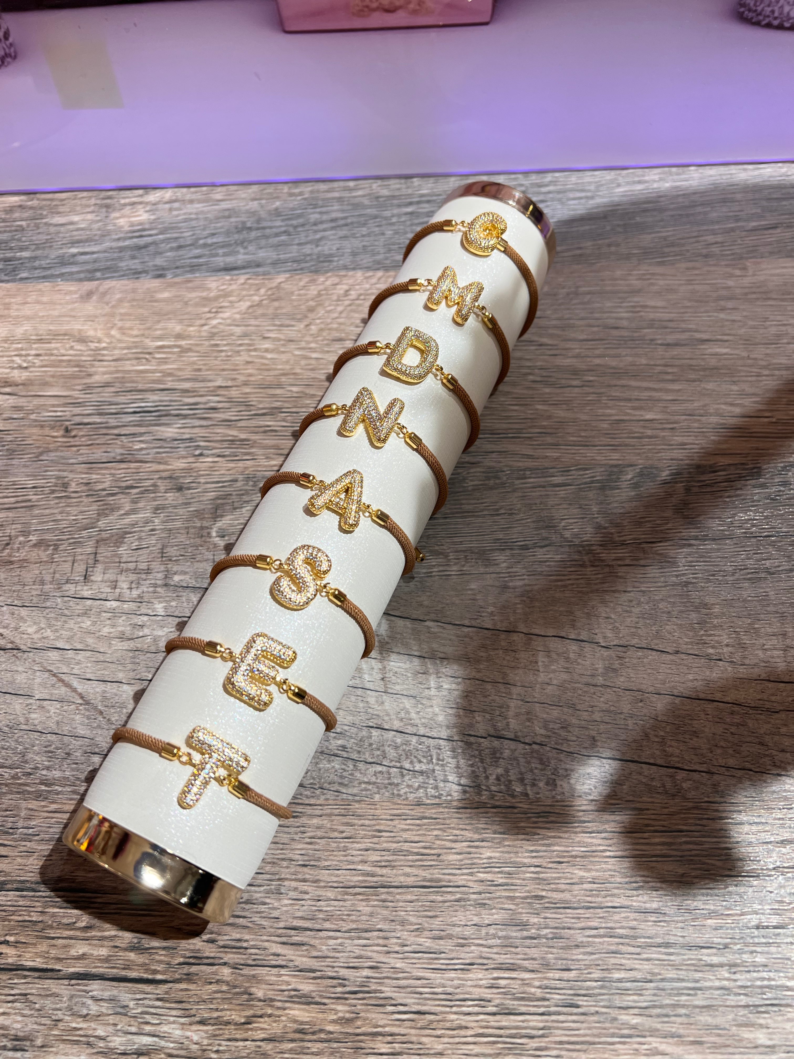 Mehrere goldfarbene Armbänder mit glitzernden Buchstaben Elementen C, M, D, N, A, S, E, und T auf einem cremefarbenen Präsentationsständer, liegend auf einer Holzoberfläche.