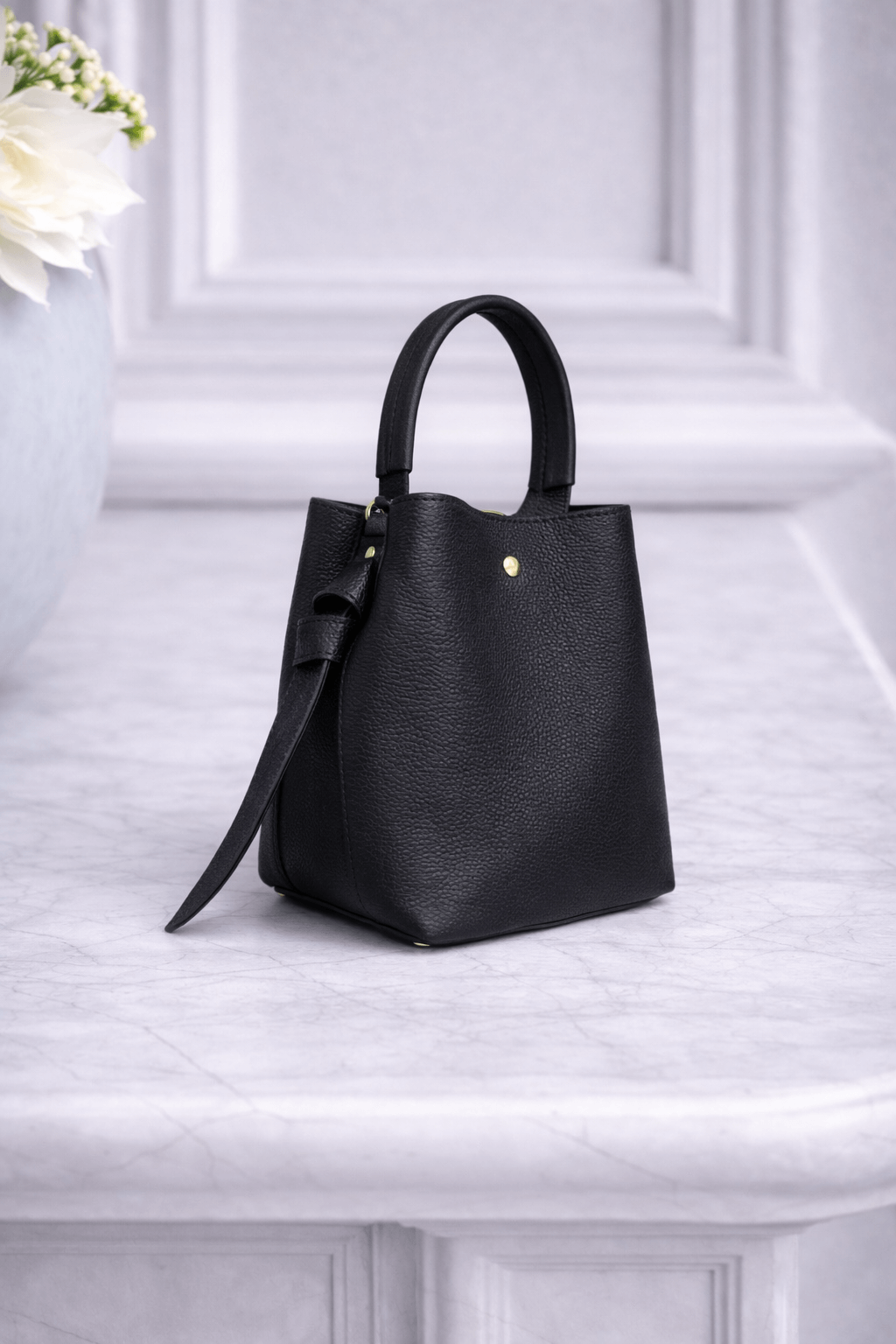 Kompakte Handtasche aus schwarzem Nappaleder mit rundem Henkel und goldfarbener Niete, stehend auf einer hellen Marmorfläche vor ruhigem Hintergrund.
