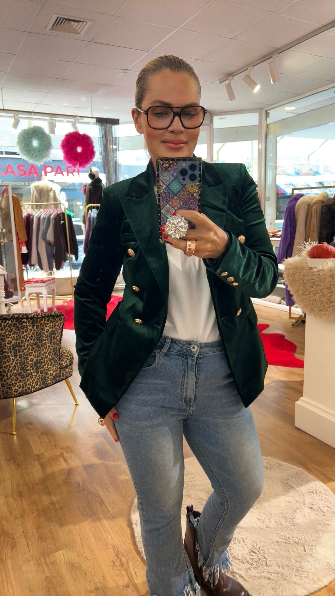Frau trägt einen grünen Samtblazer mit goldenen Knöpfen. Dazu eine Jeans mit braunen Stiefeln. Sie hält ein Handy in der Hand. Fotografiert in einer Boutique