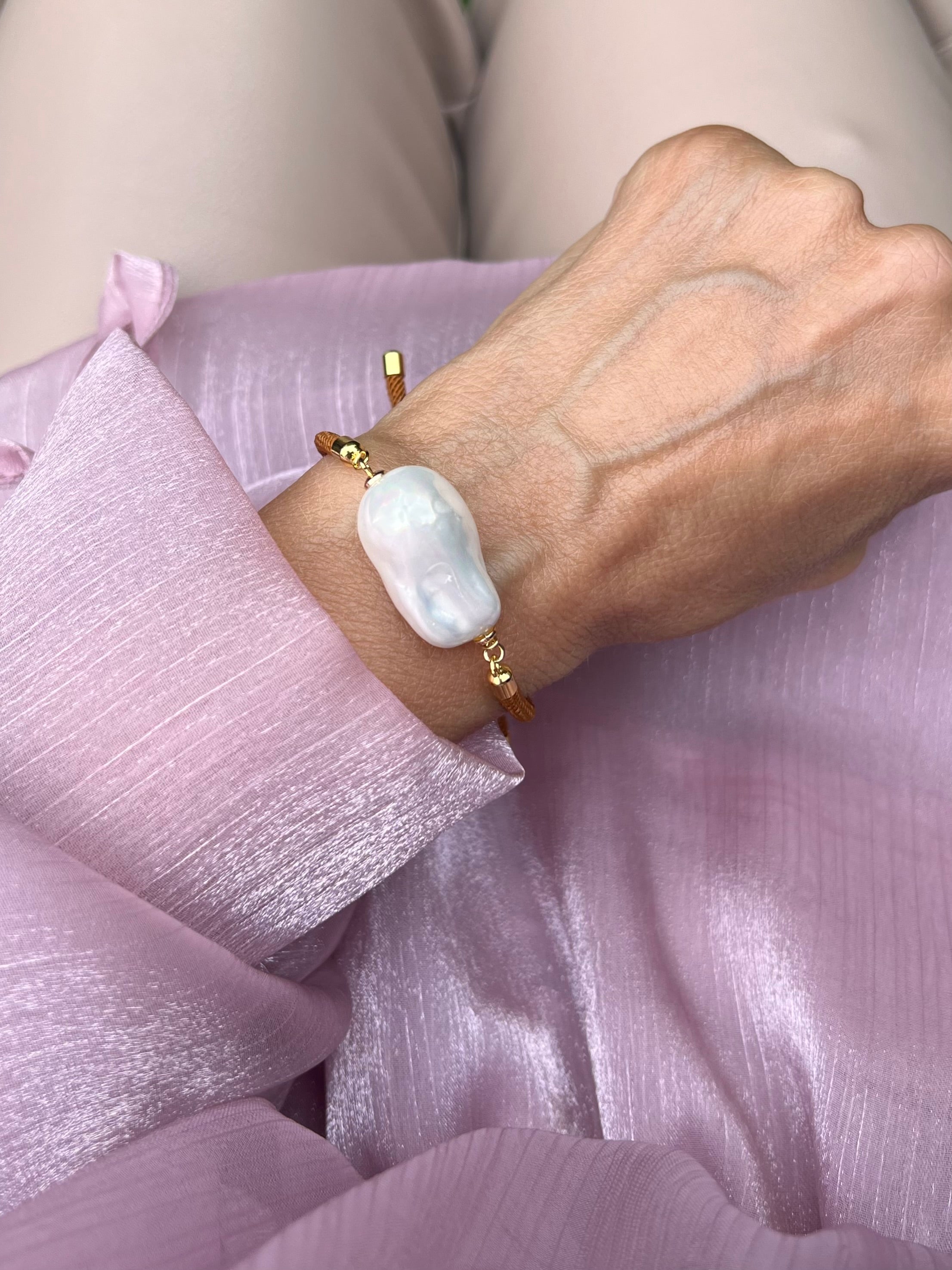 Handgelenk mit feinem braunen Armband und einer Barockperle mit goldenen Details. Auf rosa Stoff fotografiert.