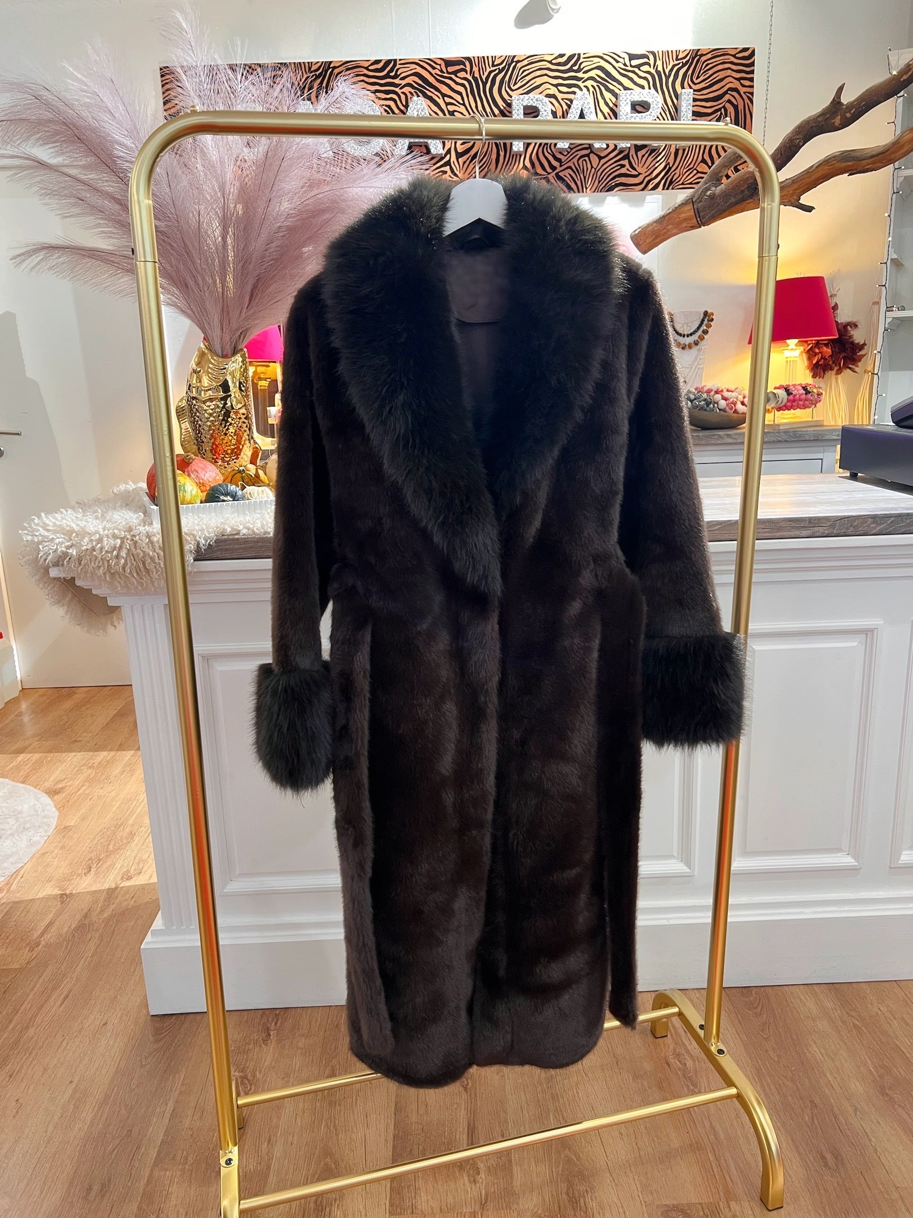 Brauner flauschiger Kunstpelzmantel hängt an einem goldenen Kleiderständer. Im Hintergrund eine weiße Theke.