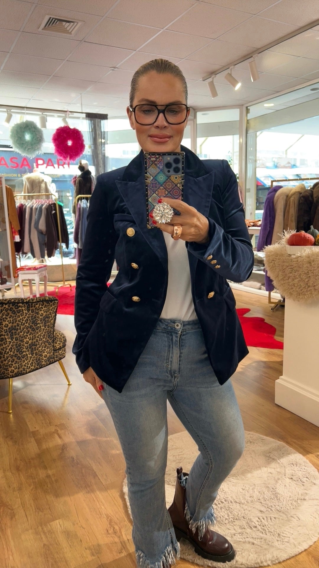 Frau trägt dunkelblauen Samtblazer mit goldenen Knöpfen. Sie hält ein Handy in der Hand, trägt eine große Brille dazu Jeanshose und Stiefel. In einer Boutique fotografiert.