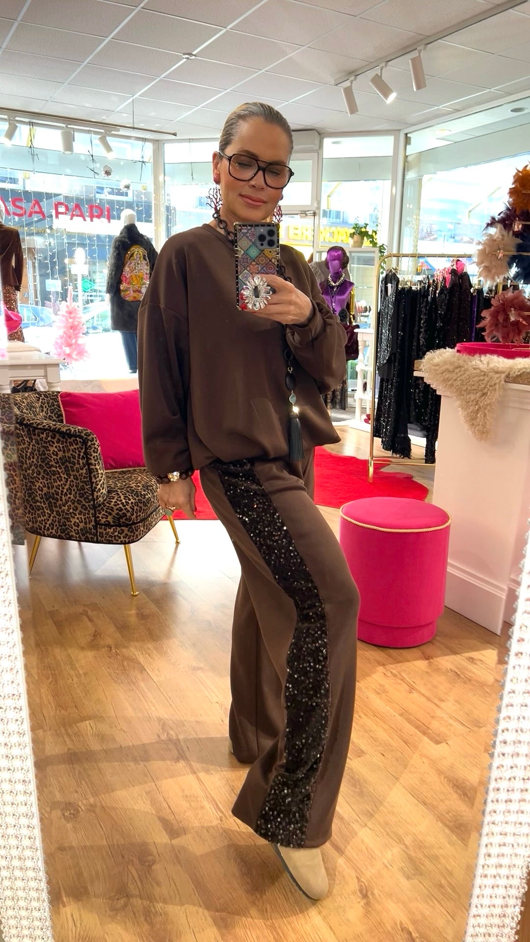 Person steht in einem braunen Zweiteiler mit funkelndem Paillettenstreifen an der Seite, in einer Boutique mit Spiegel und Dekoration.
