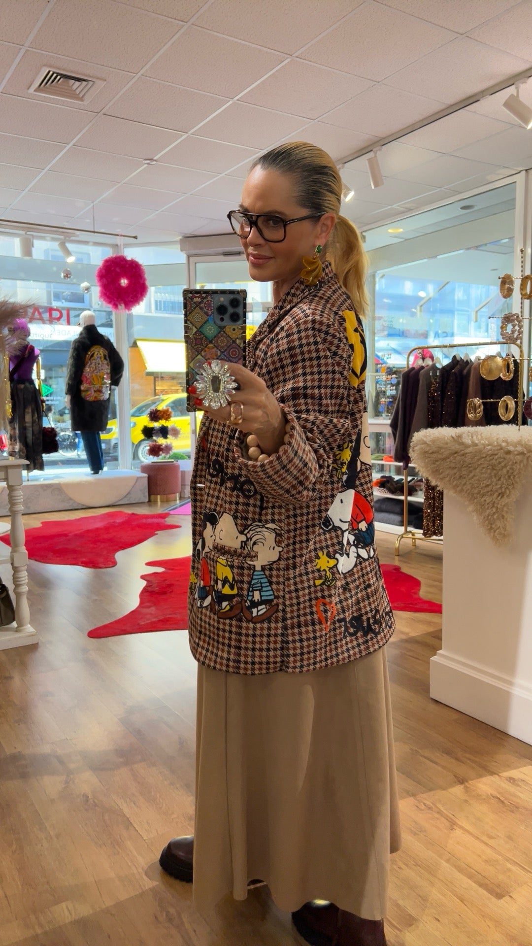 Seitliche Aufnahme derselben Person im Boutique-Store beim Spiegel-Selfie. Sichtbar ist der karierte Blazer mit großflächigen Comic-Motiven auf Rücken und Seite sowie ein langer beiger Rock oder Hose und dunkle Stiefel. Der Laden zeigt Kleidung und Accessoires im Hintergrund.