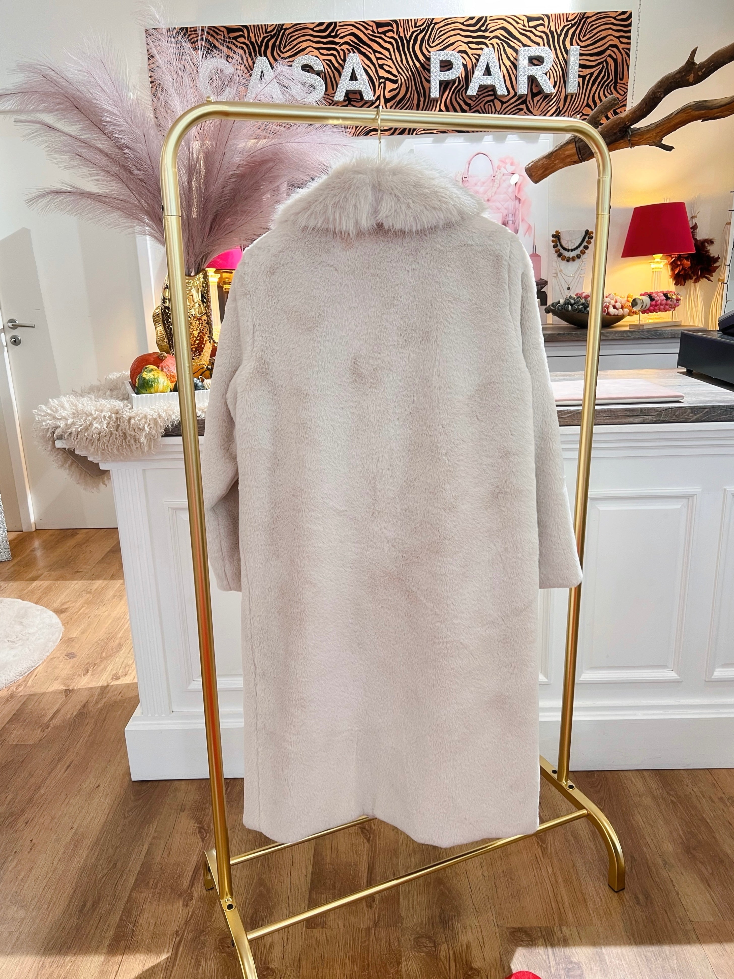 Rückansicht eines langen flauschigen Mantels in Creme hängt auf einem goldenen Kleiderständer vor einer weißen Theke.