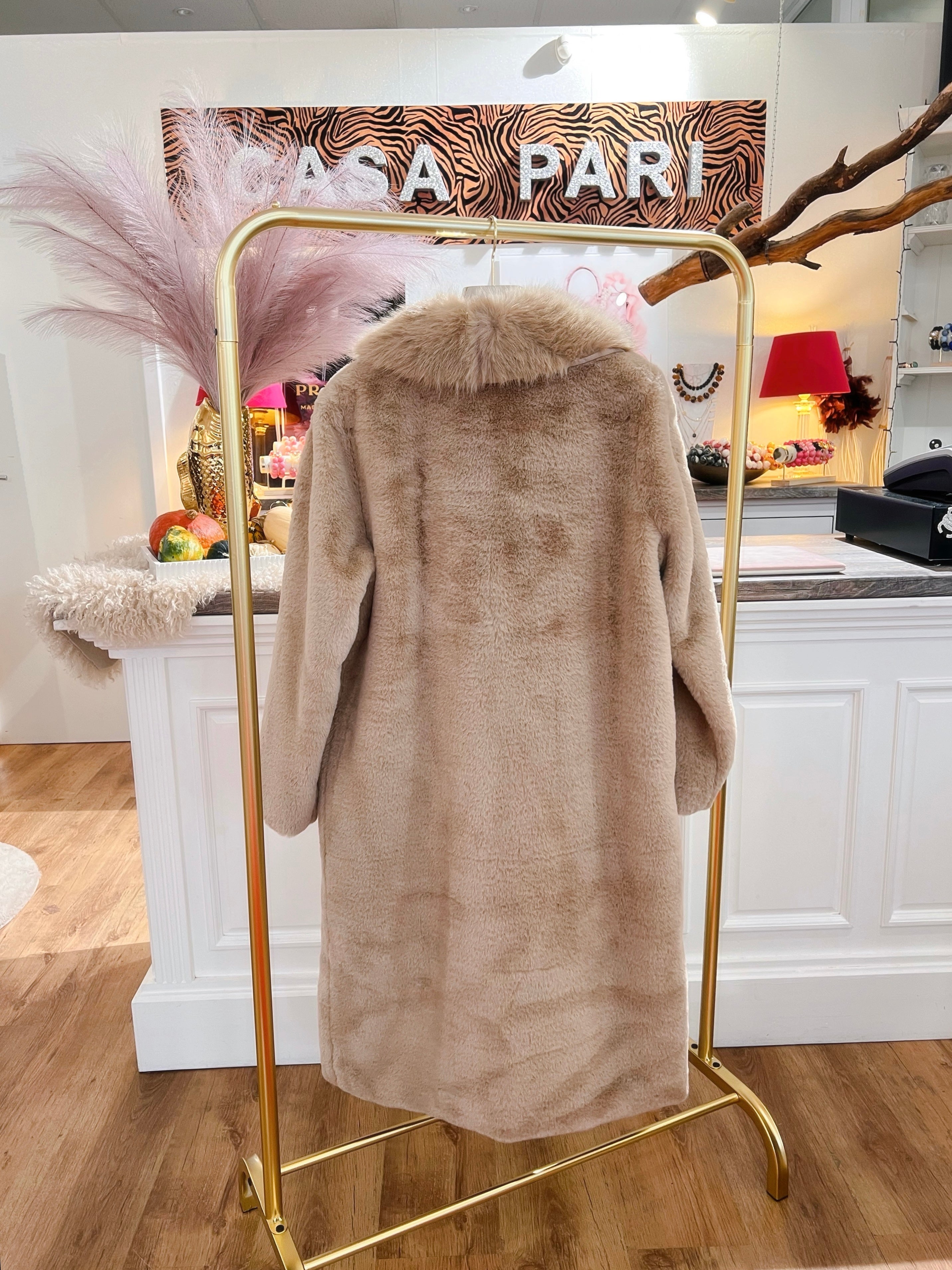 Rückenansicht eines langen, flauschigen Mantel in Beige auf einem goldenen Kleiderständer vor einer weißen Theke.