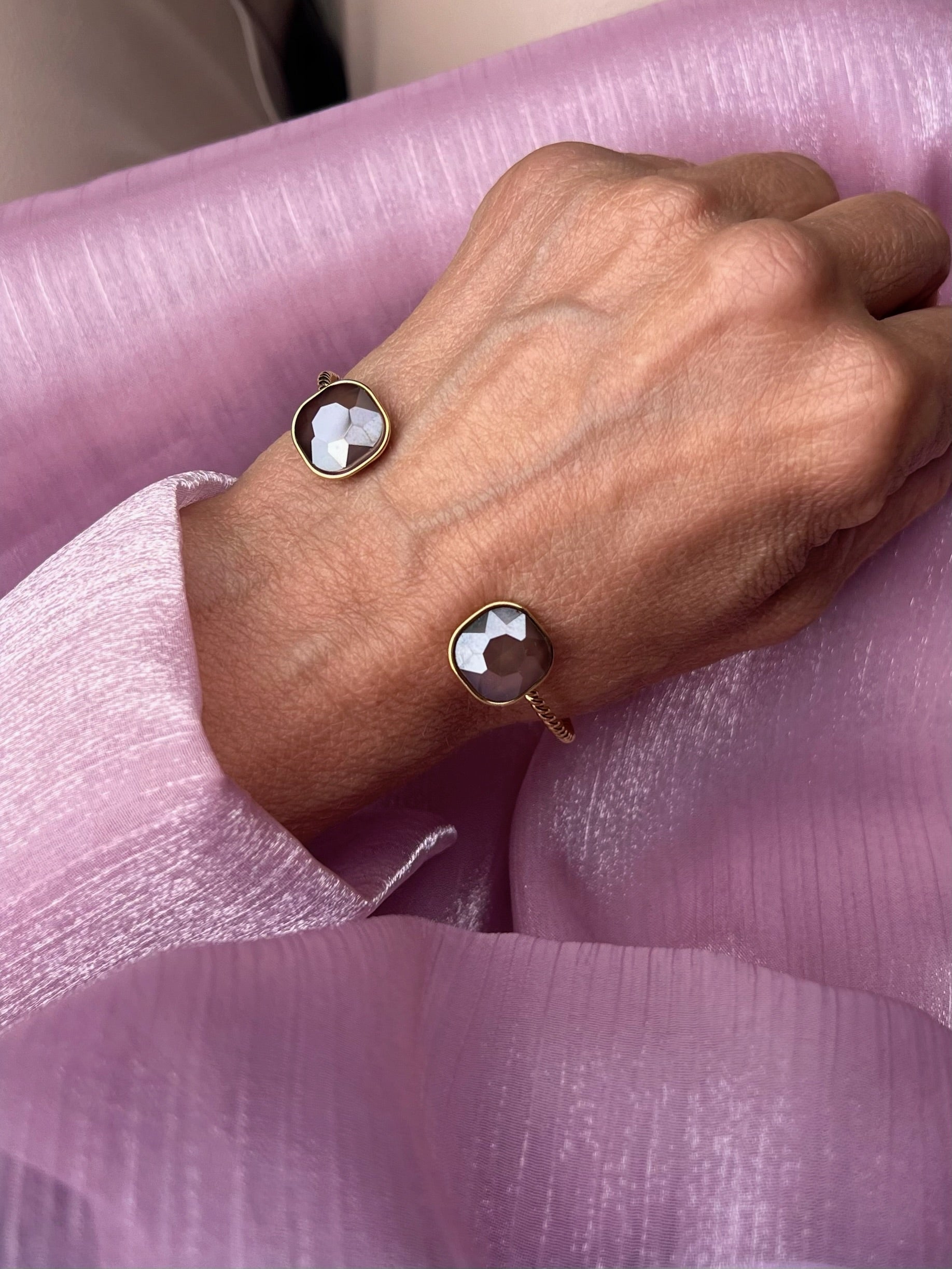 Armreifen in Gold mit Stein in Taupe, fotografiert an einem Handgelenk auf Rosa Stoff.