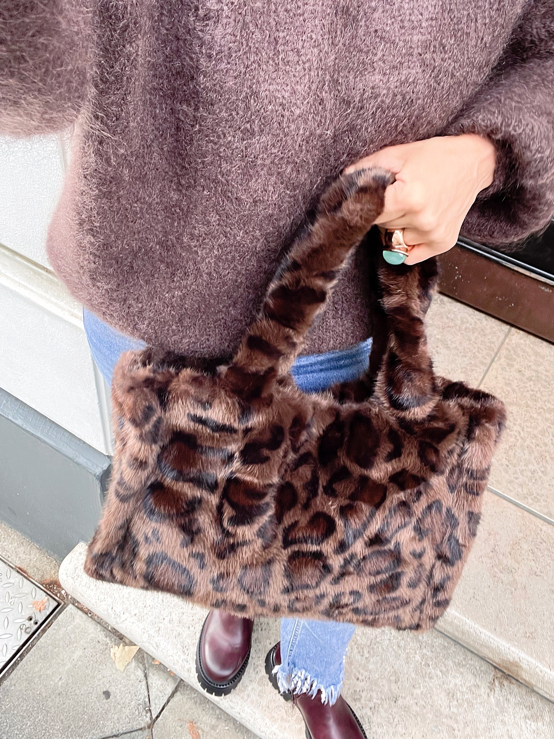 Frau trägt Leo Fell Plüschtasche in der Hand mit einem großen Ring. Dazu einen braunen Mohairpulli dazu. Jeans fotografiert an einer Türe.