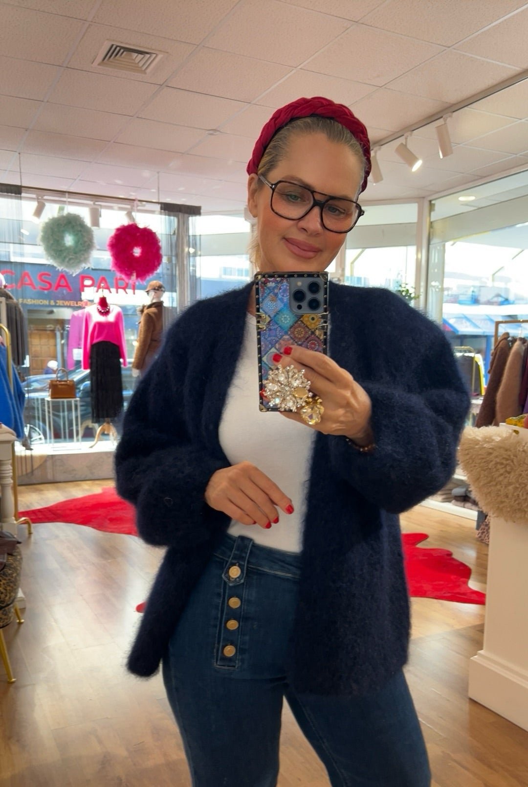 Frau trägt flauschige Strickjacke in Dunkelblau, kombiniert mit weißem Shirt und dunkelblauer Jeans mit Knopfleiste. Im Hintergrund bunte Mode und rote Akzente.