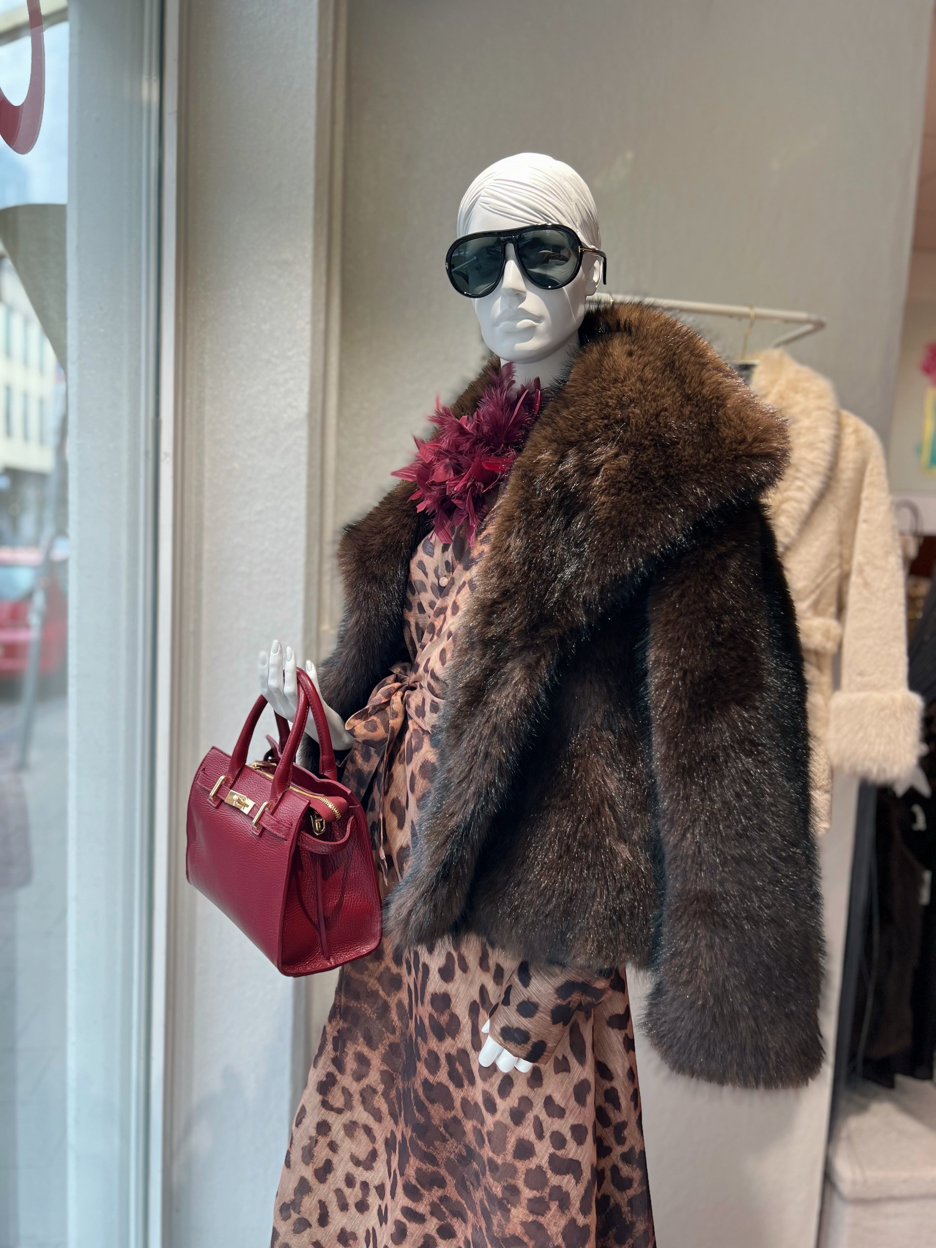 Schaufensterpuppe trägt braune Pelzjacke ein Leo Kleid und eine Handtasche in Rot, mit Sonnenbrille. Am Kragen eine rote Federbrosche.