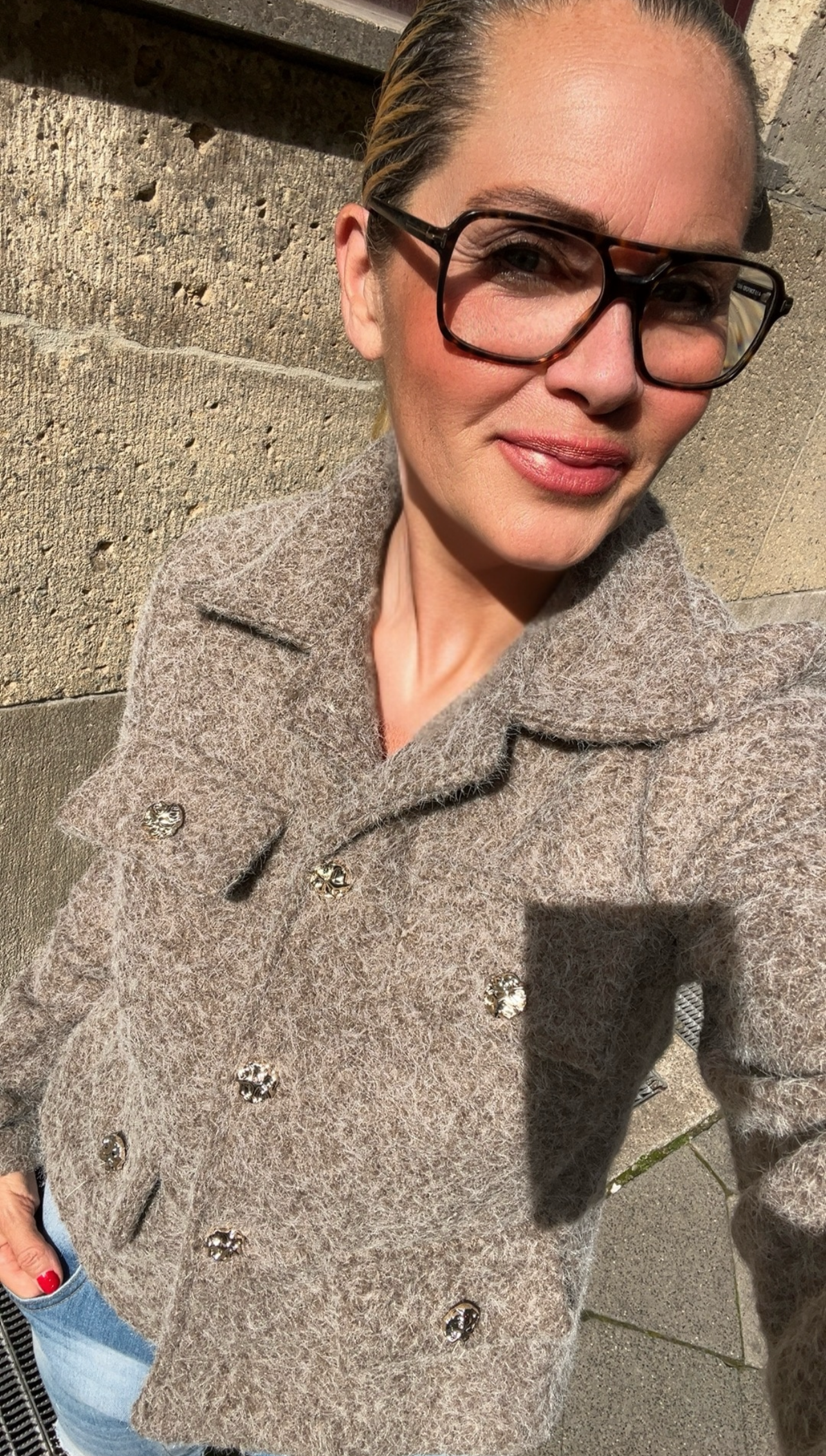 Frau steht an Wand trägt beigen Blazer mit goldenen Knöpfen und trägt große Brille.