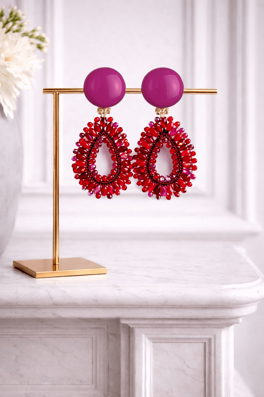 Garne-Ohrringe in Rot mit pinkem Cabochon und tropfenförmigem Anhänger aus facettierten Glasperlen an goldfarbenem Schmuckständer auf hellem Marmortisch.