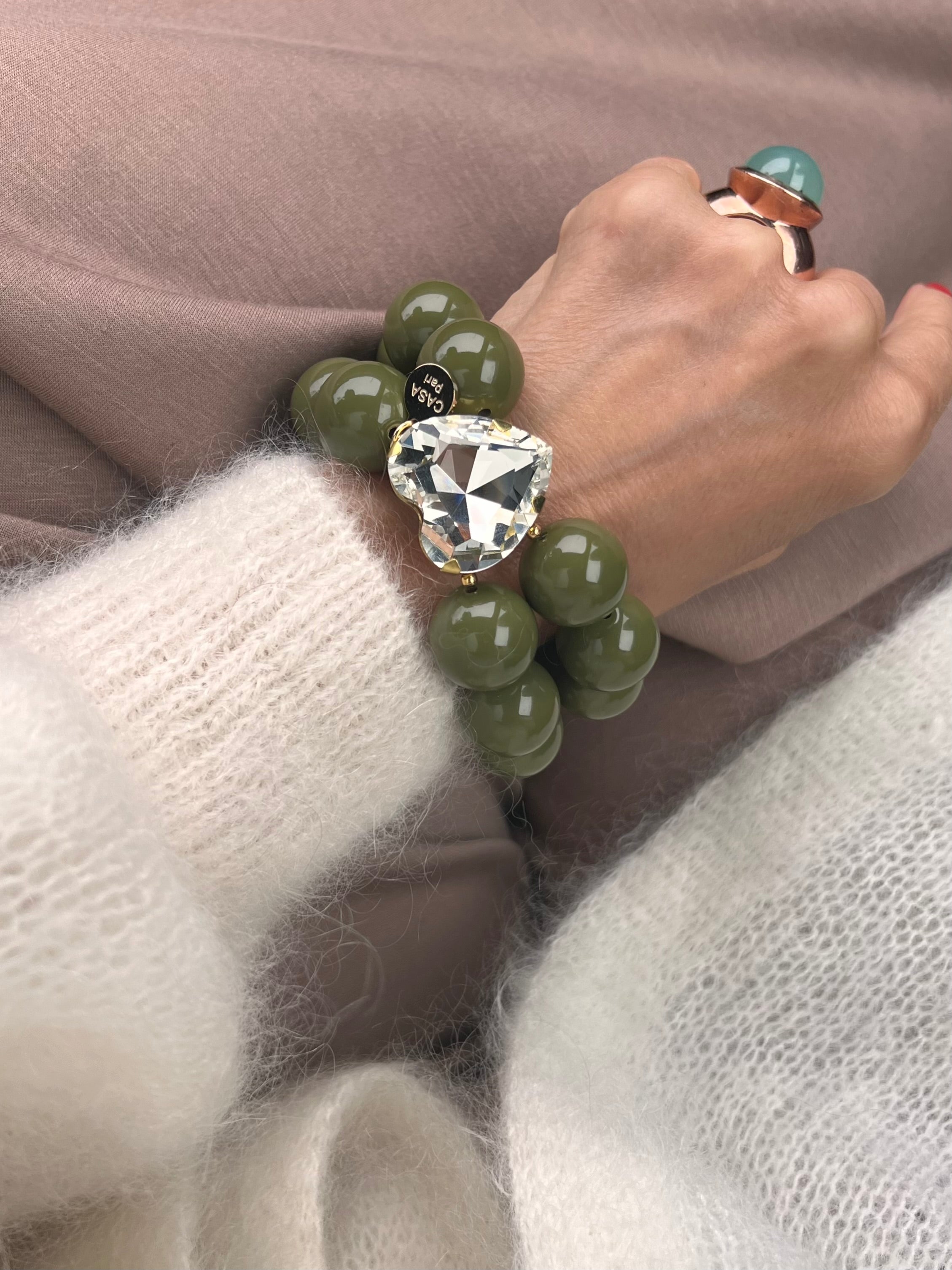 Armband in Olivenfarben mit großem Herzschmuckstein, und großem Ring. Am Handgelenk auf beigem Stoff fotografiert.