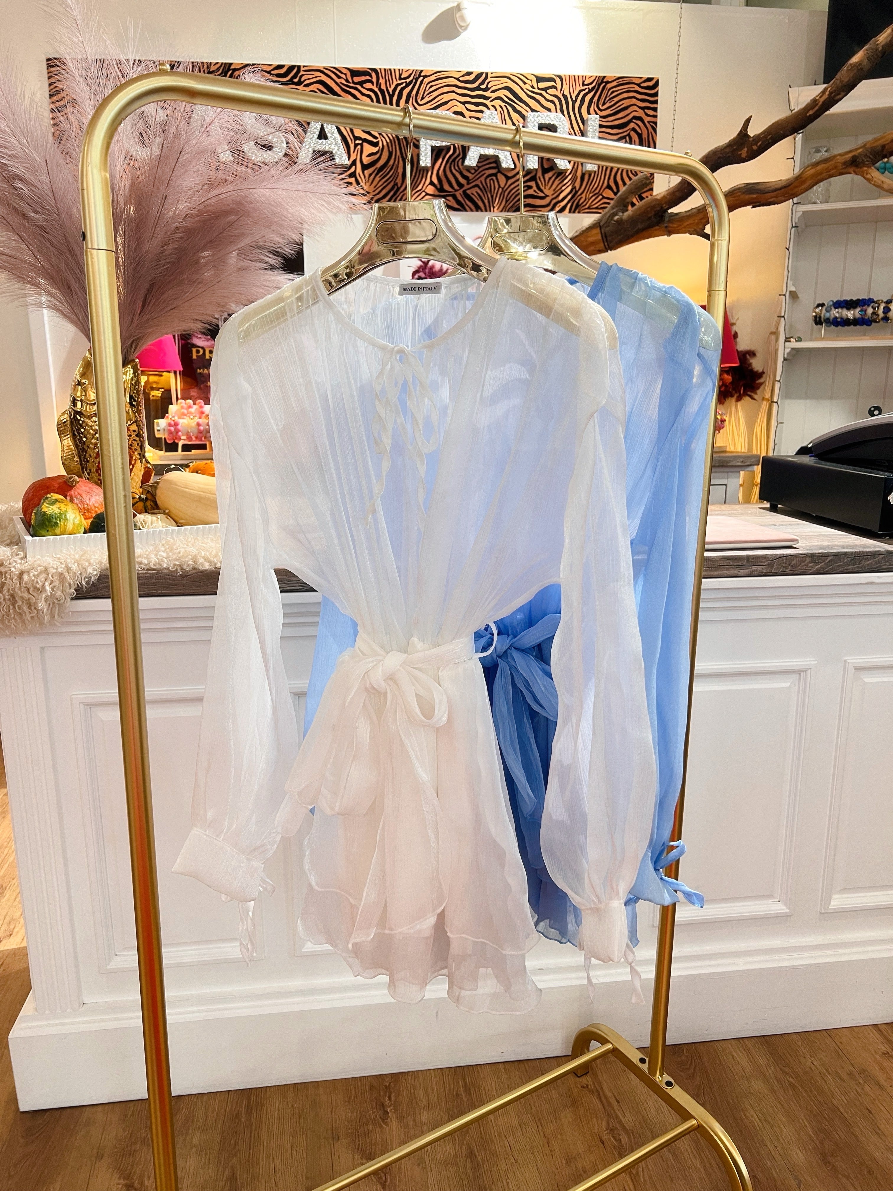 Mehrere transparente Blusen in Weiß, und Hellblau hängen auf goldenen Kleiderbügeln an einem goldenen Kleiderständer vor weißen Theke im Laden.