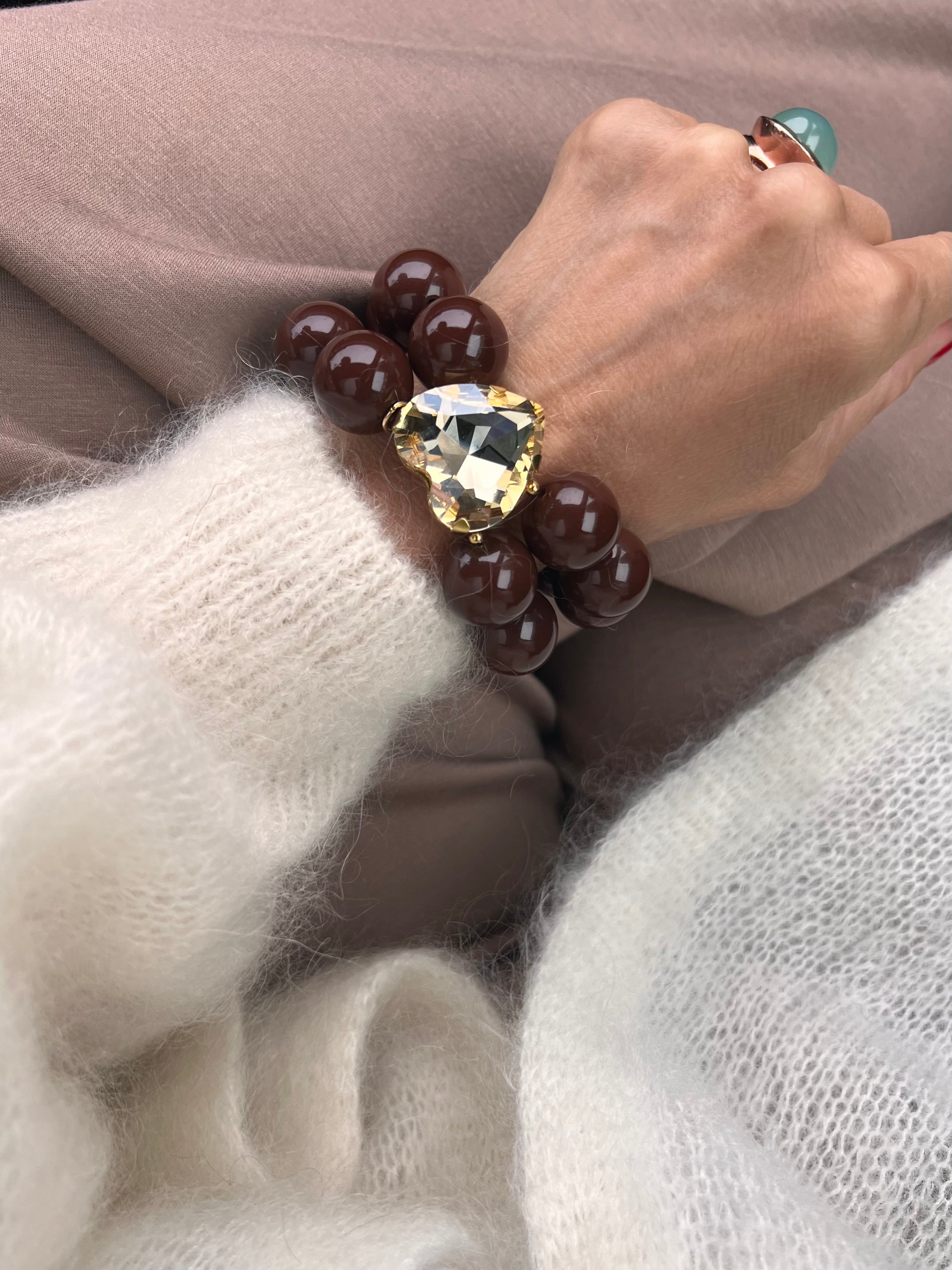 Armband in Dunkelbraun mit großem Herzschmuckstein, und großem Ring. Am Handgelenk auf beigem Stoff fotografiert.