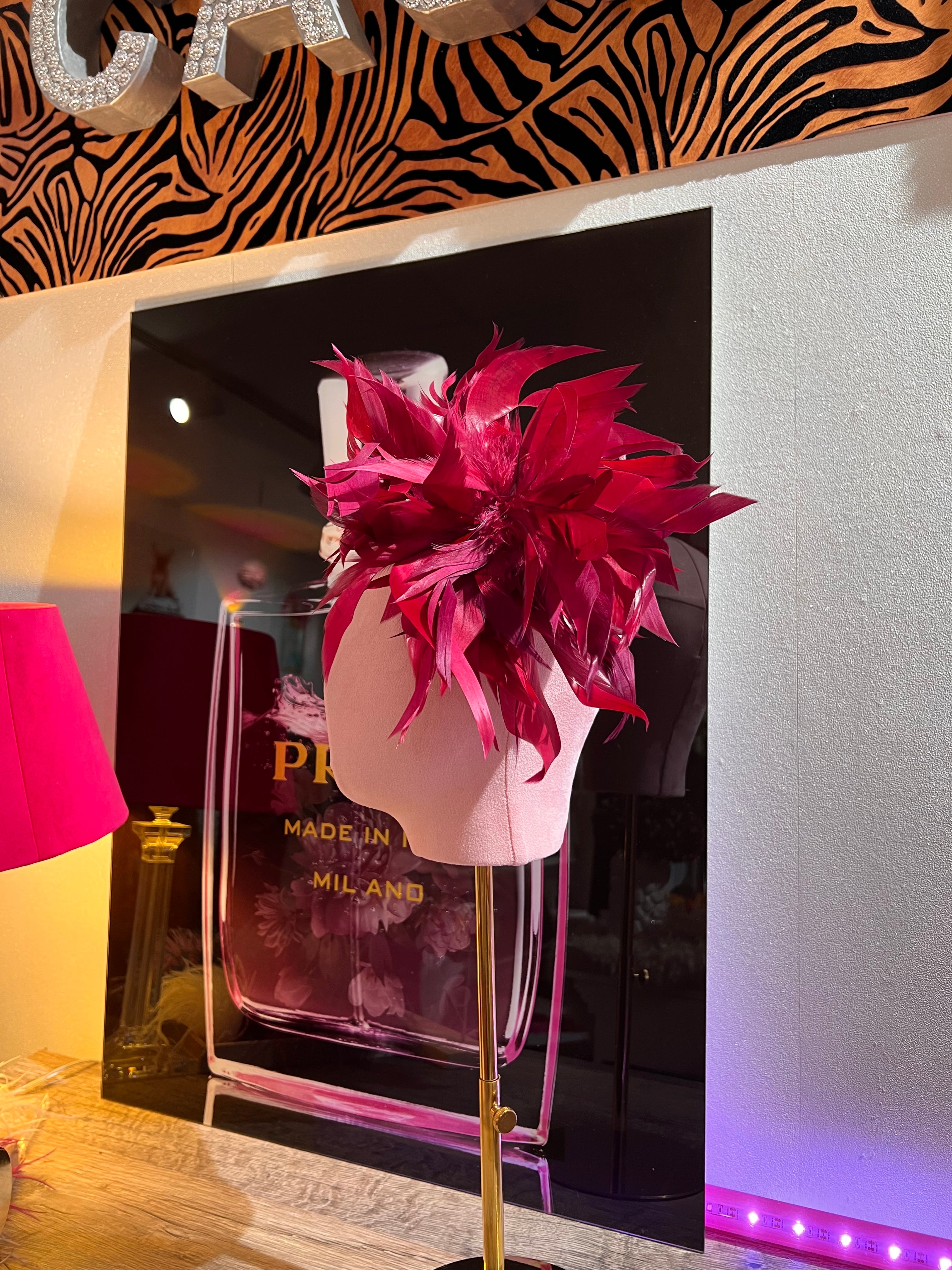 Federbrosche mit großer Blüte aus roten bordeaux farbenen Federn, befestigt auf einer rosa Stoffpuppe vor dekorativen Hintergrund mit Parfumflakon Motiv.