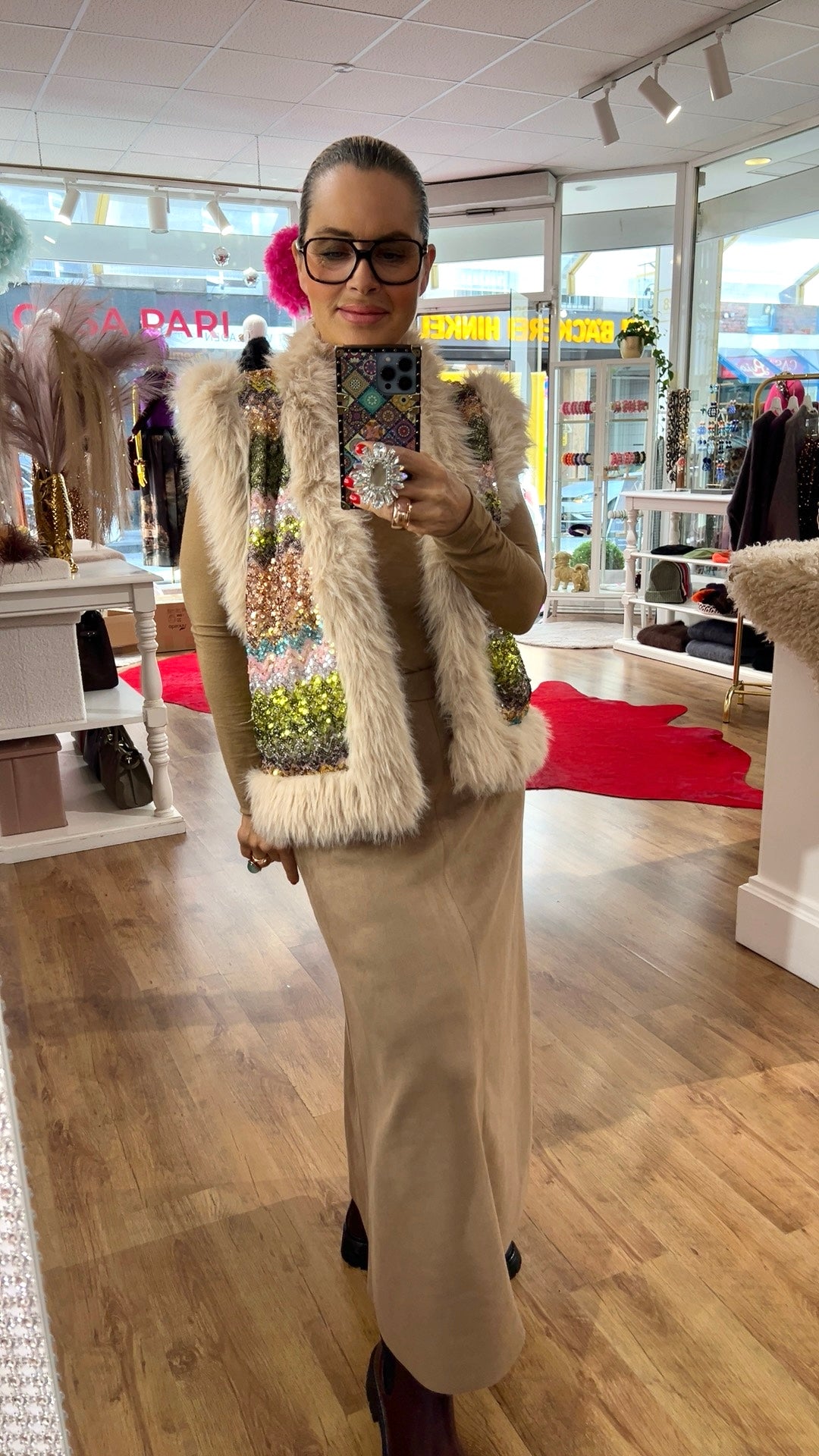Person steht in einem Boutique-Innenraum und fotografiert sich im Spiegel. Sie trägt eine cremefarbene Kunstfell-Weste mit funkelnden, mehrfarbigen Pailletten auf der Vorderseite über einem beigefarbenen, langärmligen Oberteil und einem langen beigen Rock. Die Person hält ein Smartphone mit dekorativem Pop-Grip. Im Hintergrund sind Regale mit Schmuck und Accessoires sowie Dekorationen zu sehen.