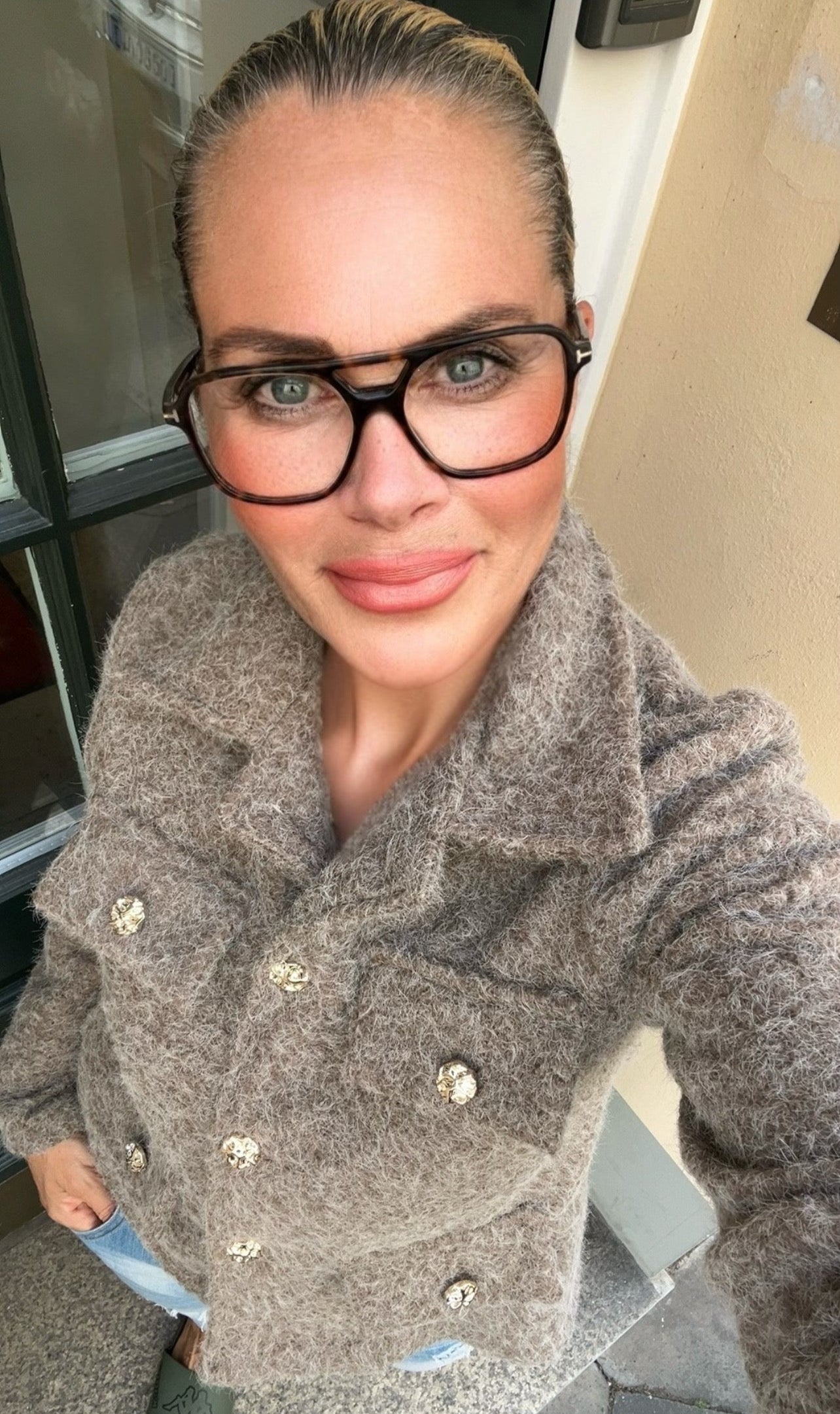 Frau steht in Türe trägt taupe farbene Jacke mit goldenen Knöpfen und großer Brille.