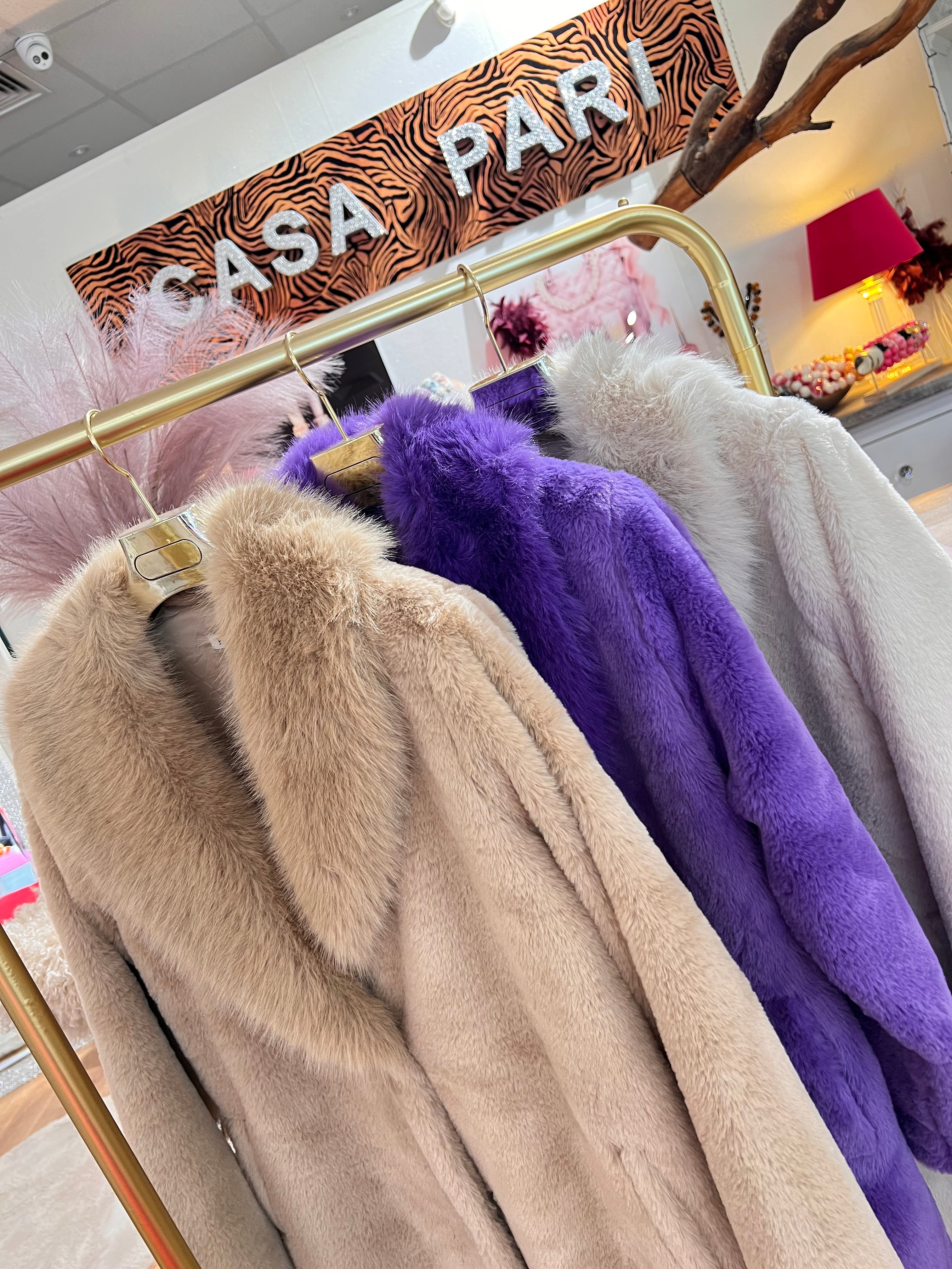 Mehrere flauschige Mäntel in Beige, Violett und Creme hängen auf einem goldenen Kleiderständer im Geschäft mit dekorativen Elementen im Hintergrund.