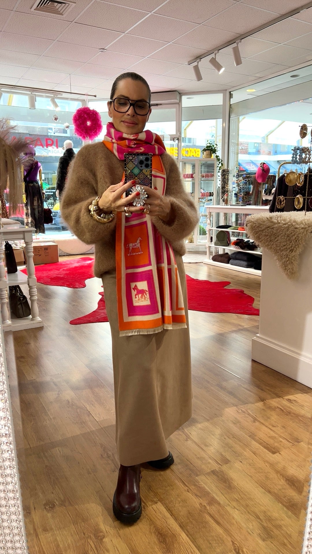 Person steht in einem Boutique-Innenraum und fotografiert sich im Spiegel. Sie trägt einen beigen, flauschigen Pullover, einen langen beigefarbenen Rock und dunkle Boots. Um den Hals liegt ein bunter Schal in Pink-, Orange- und Weißtönen mit Pferdemotiven. Am Handgelenk sind mehrere Armbänder zu sehen. Im Hintergrund befinden sich Regale mit Schmuck und Accessoires sowie Dekorationselemente.