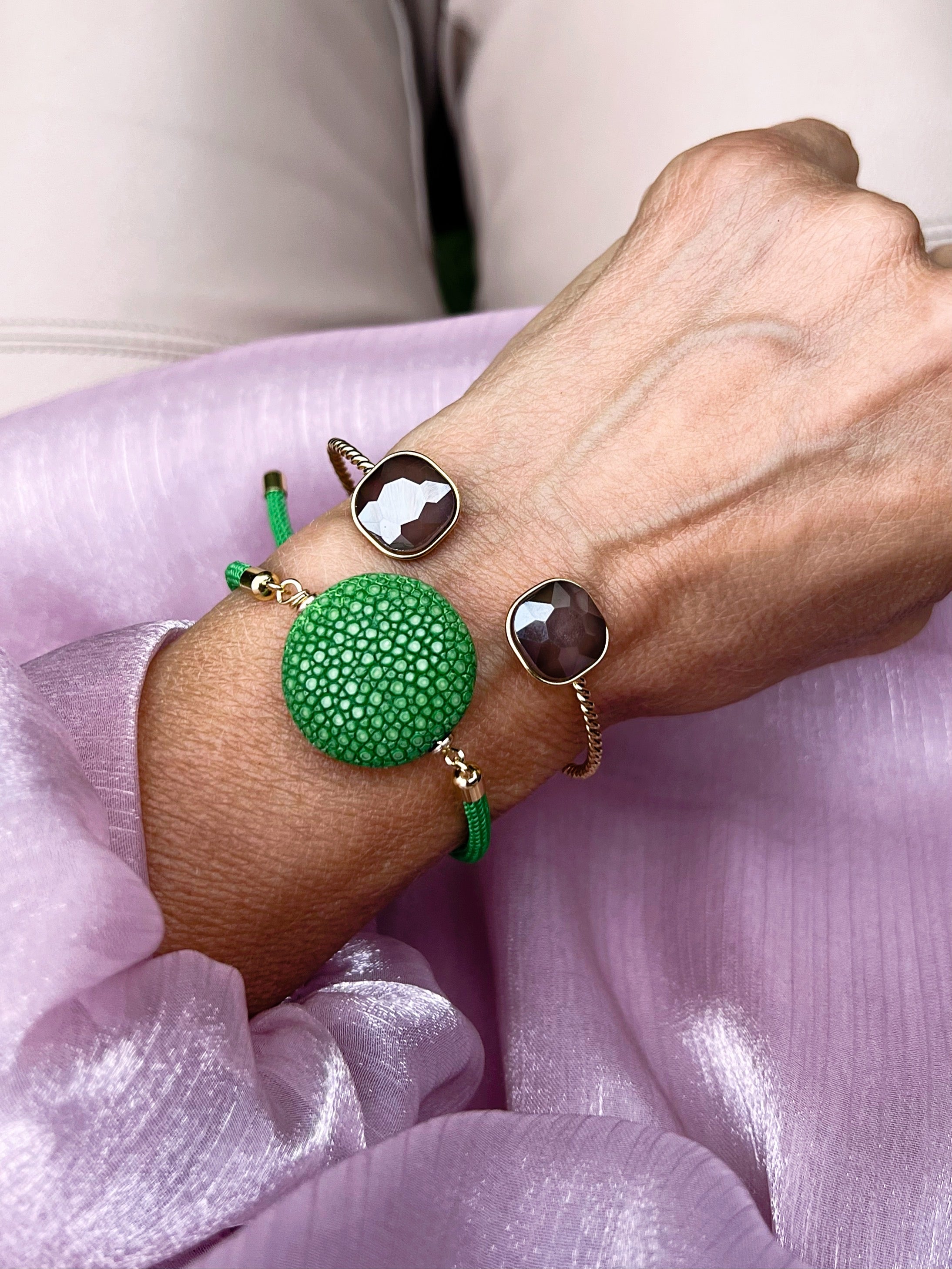 Ein grünes Armband mit grüner Perle dazu ein goldener Armreif mit Glasstein in Taupe. An einem Handgelenk auf rosa Stoff fotografiert.