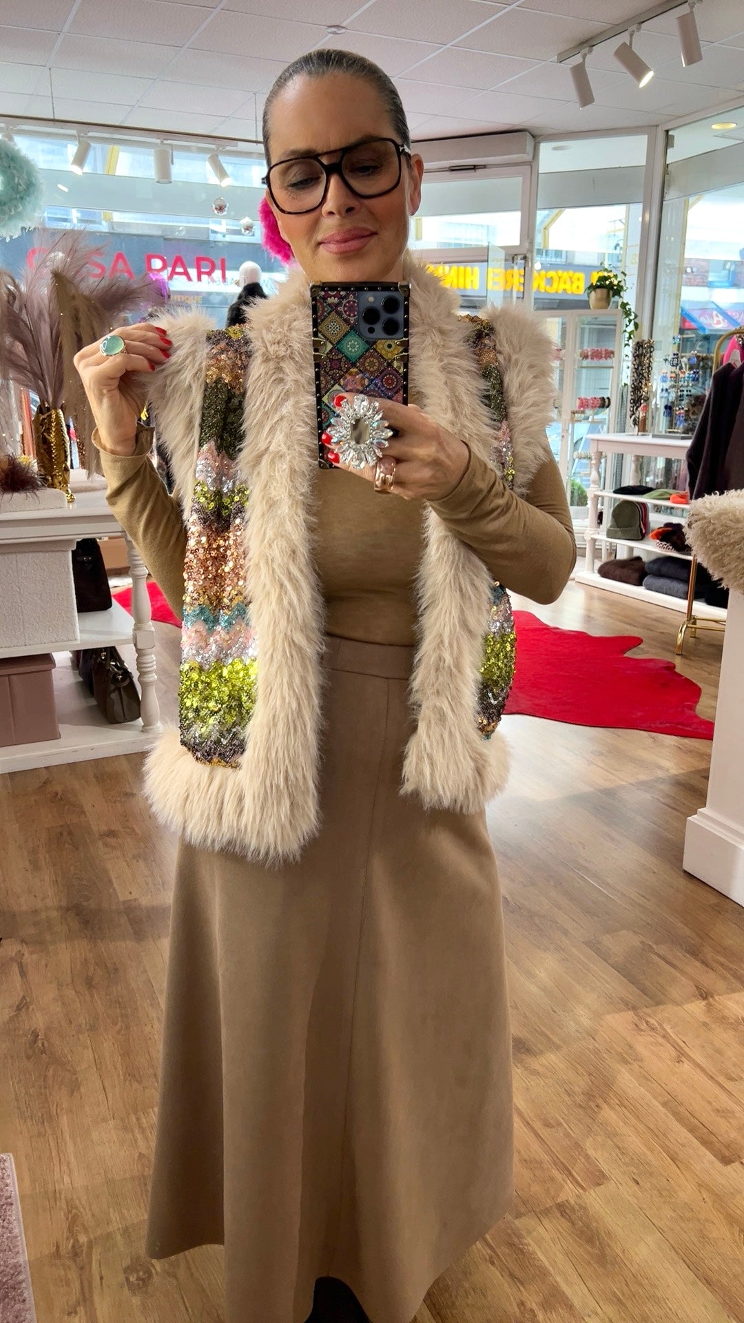 Person steht in einem Boutique-Innenraum und fotografiert sich im Spiegel. Sie trägt eine cremefarbene Kunstfell-Weste mit funkelnden, mehrfarbigen Pailletten auf der Vorderseite über einem beigefarbenen, langärmligen Oberteil und einem langen beigen Rock. Die Person hält ein Smartphone mit dekorativem Pop-Grip. Im Hintergrund sind Regale mit Schmuck und Accessoires sowie Dekorationen zu sehen.