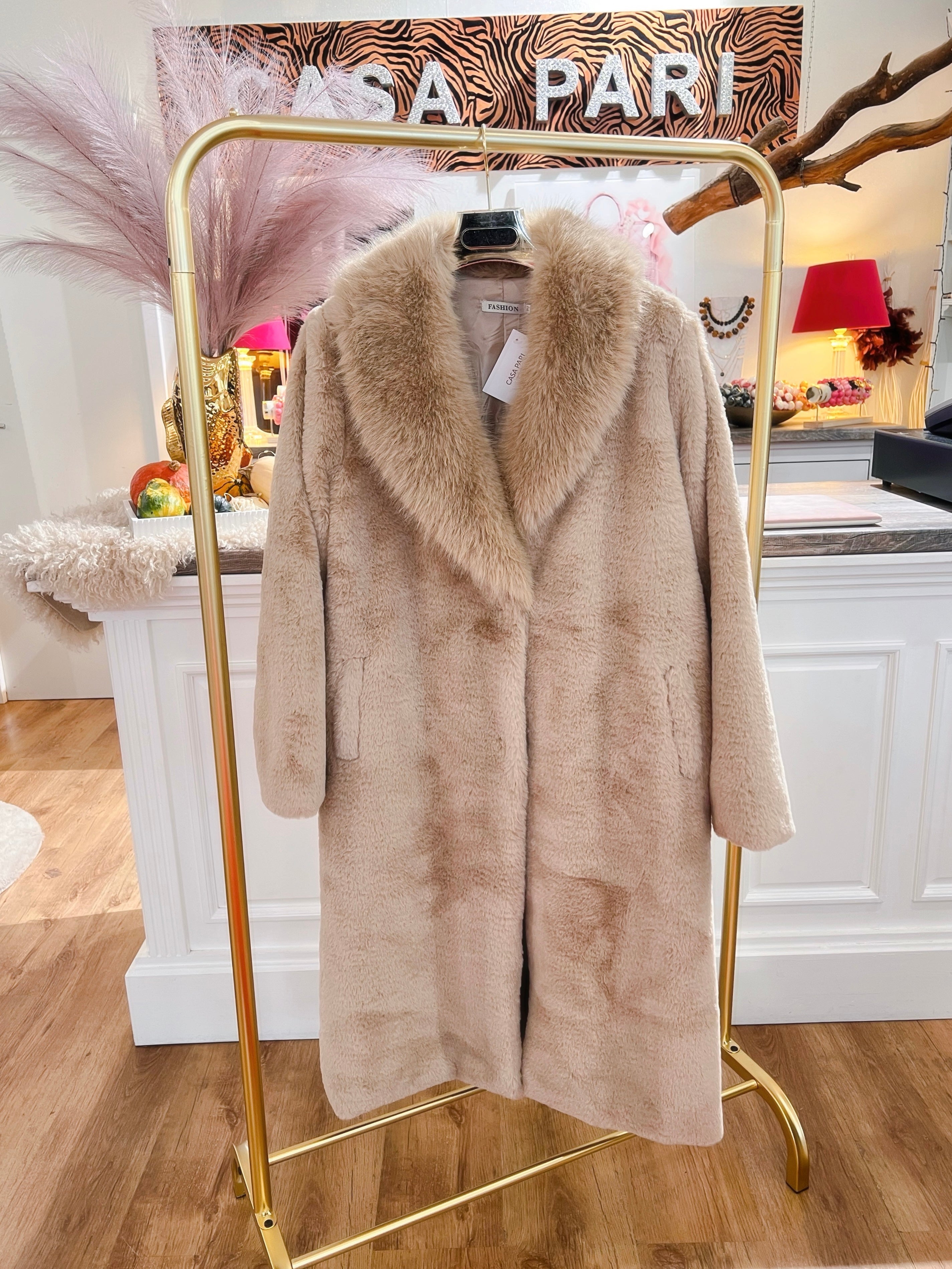 Langer, flauschiger Mantel in Beige hängt auf einem goldenen Kleiderständer vor einer weißen Theke.