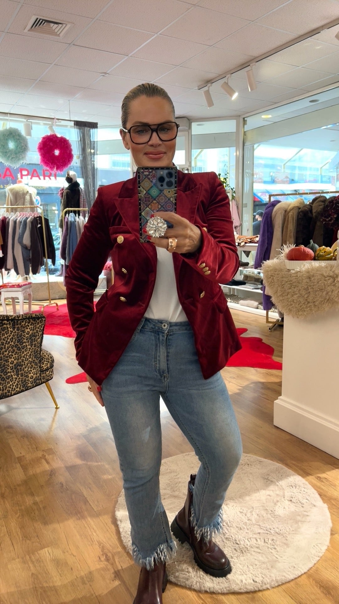 Frau trägt weinroten Samtblazer mit goldenen Knöpfen. Dazu eine Jeans und braune Boots. Sie hält ein Hand in der Hand. Fotografiere in einer Boutique.