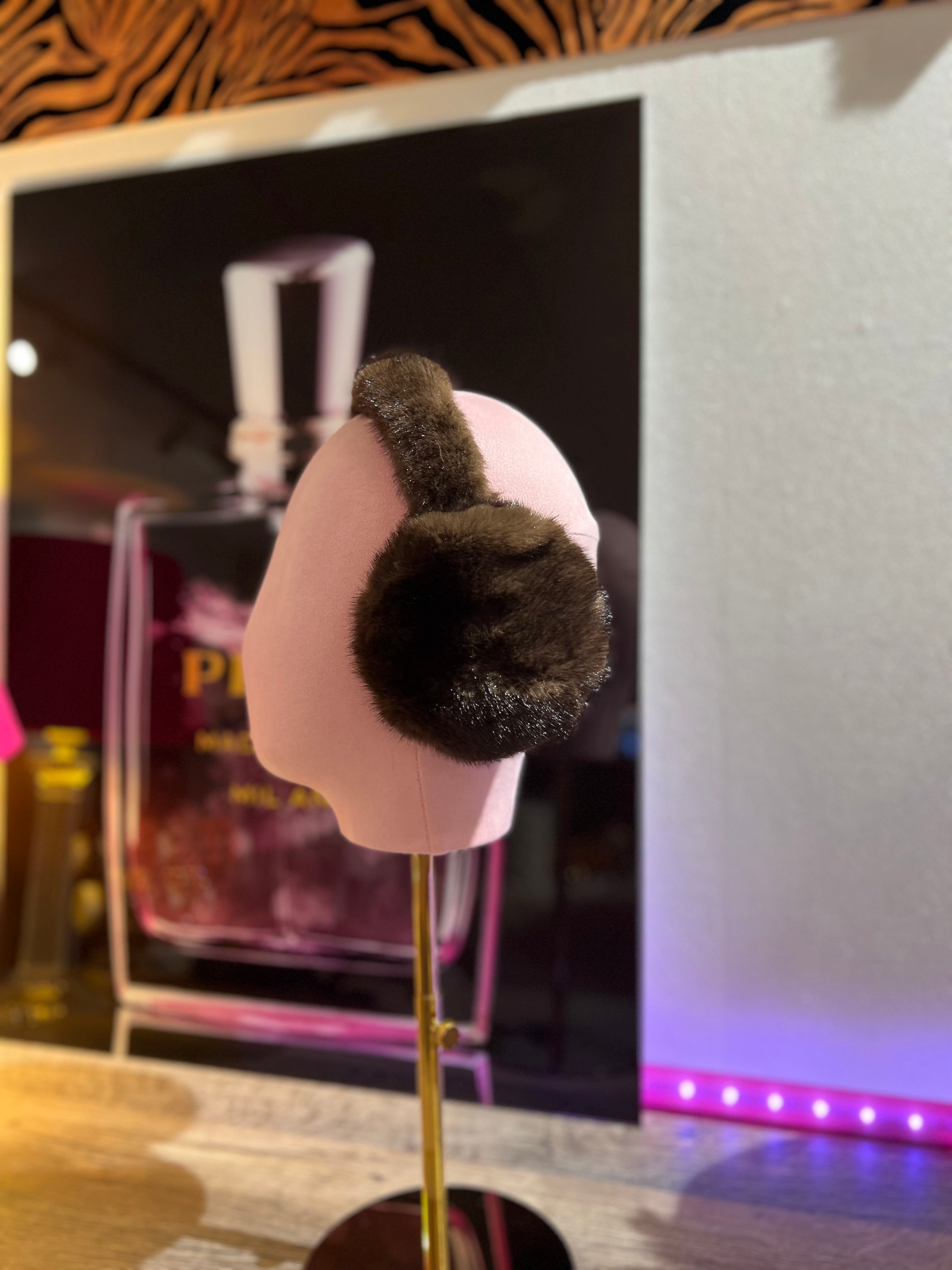 Braune, flauschige Ohrwärmer auf rosafarbene Präsentation Büste, im Hintergrund unscharf ein Parfumflakon und warme Beleuchtung.