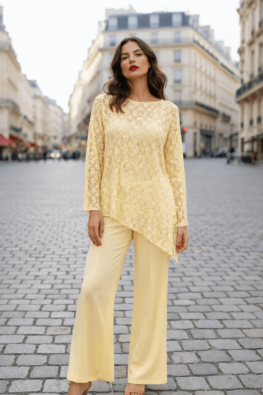 Frau steht auf einer Pariser Straße mit Kopfsteinpflaster und hellen Altbauten im Hintergrund. Sie trägt ein vanillefarbenes, asymmetrisches Spitzenoberteil mit langen Ärmeln und eine weite, hellgelbe Hose.