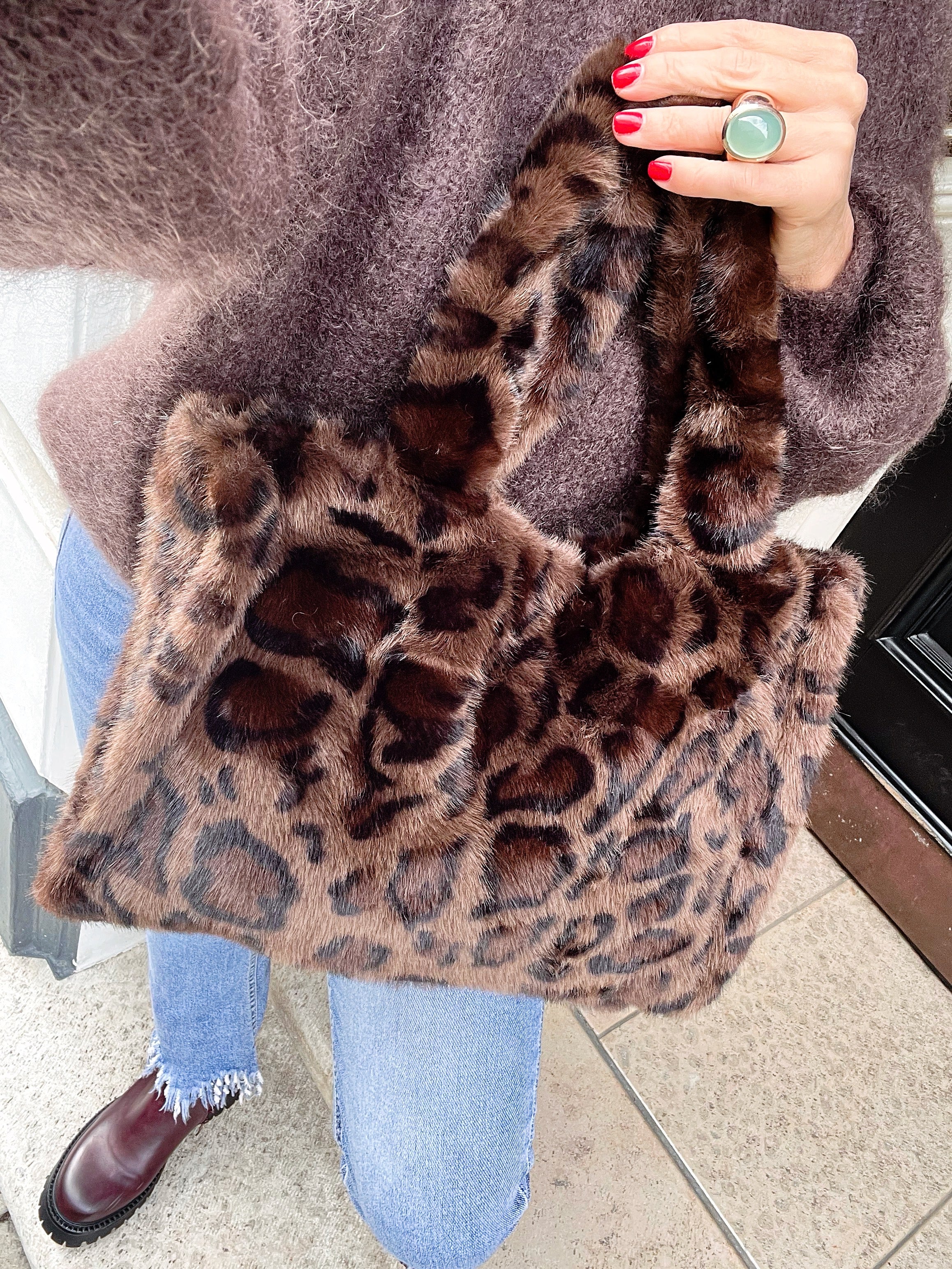 Frau trägt braune Leo Plüschtasche in der Hand mit rotem lackierten Nägeln und großem Ring. Dazu eine Jeans, brauner Mohairpulli. Fotografiert an einer Türe